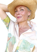 Peut inclure: Une femme porte un chapeau de paille et une chemise boutonnée de couleur claire avec un motif de paysage. Elle porte également des boucles d'oreilles carrées et colorées avec un motif tissé.