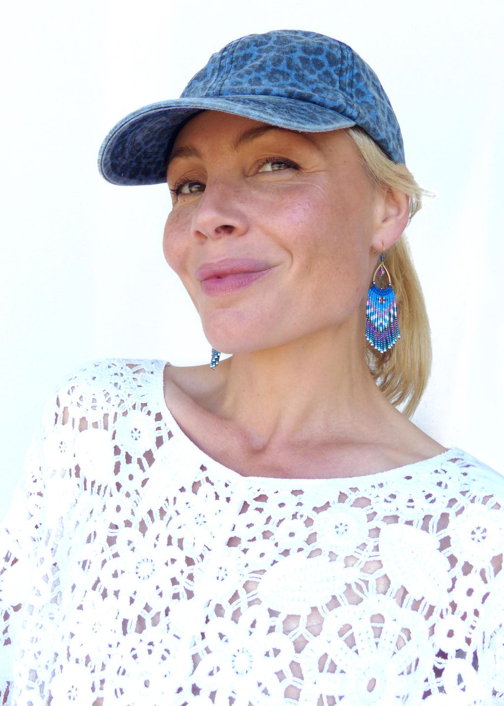 Peut inclure: Casquette de baseball en jean bleu avec un motif léopard. La photo présente des boucles d'oreilles en perles avec des franges bleues, violettes et blanches. La personne porte un haut en dentelle blanc.