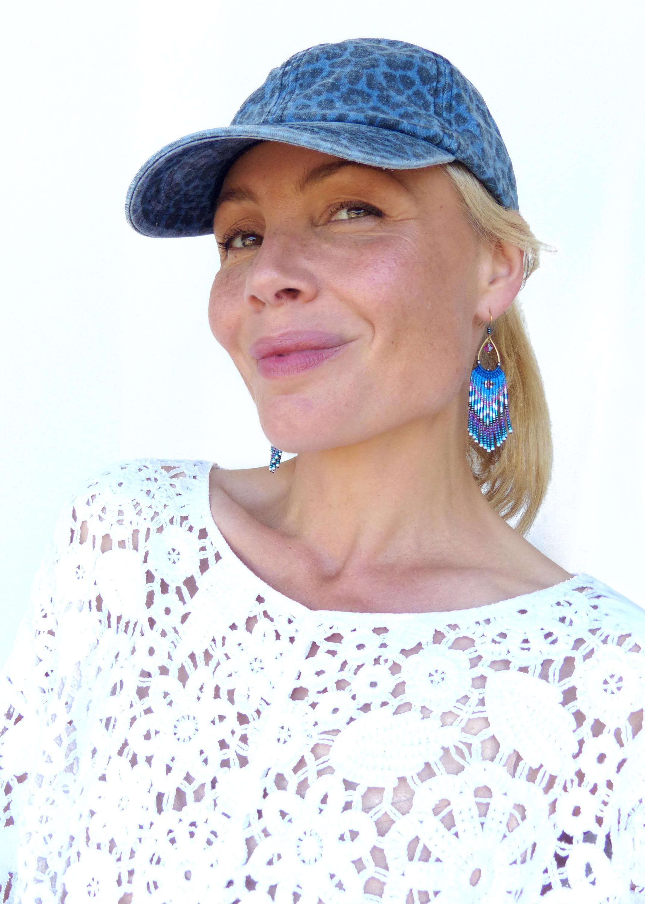 Peut inclure: Casquette de baseball en jean bleu avec un motif léopard. La photo présente des boucles d'oreilles en perles avec des franges bleues, violettes et blanches. La personne porte un haut en dentelle blanc.
