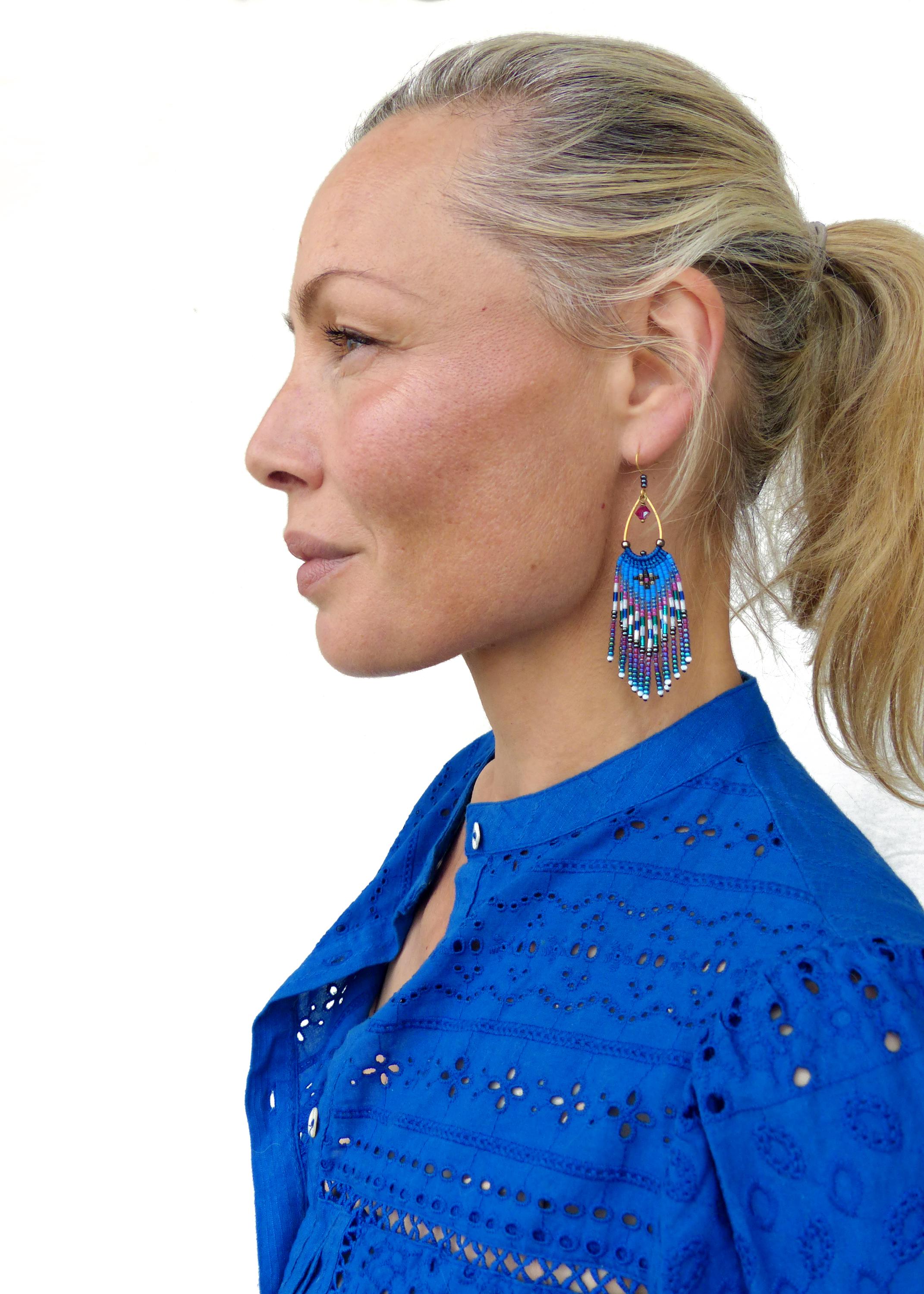 Peut inclure: Boucles d'oreilles pendantes dorées avec des franges de perles bleues, violettes et blanches. Les boucles d'oreilles ont une forme de goutte d'eau et une petite perle rouge. La femme porte une chemise bleue.