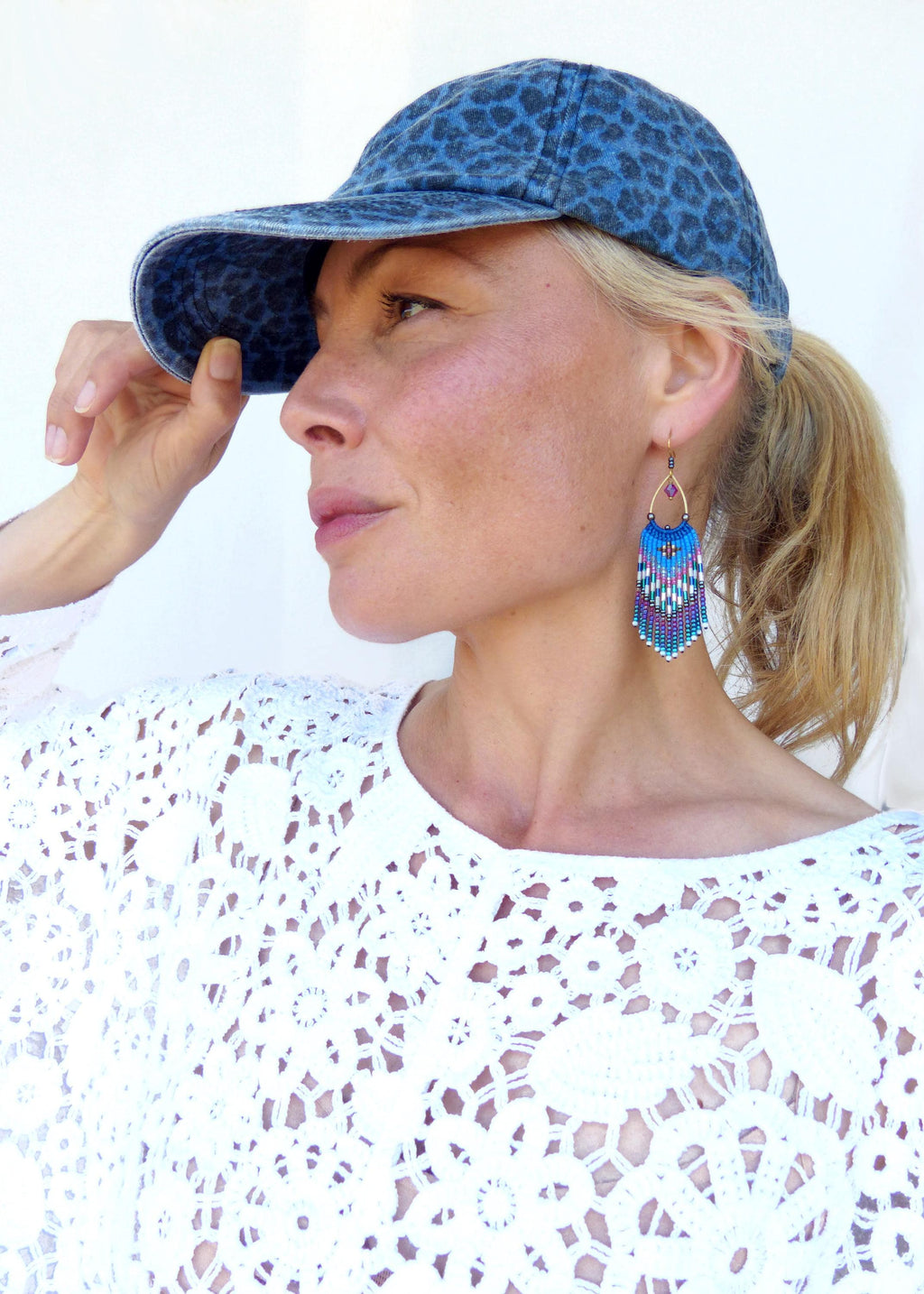 Peut inclure: Une personne porte une casquette de baseball en jean avec un imprimé léopard bleu. La personne porte également un haut en dentelle blanche et des boucles d'oreilles à franges bleues, violettes et blanches.