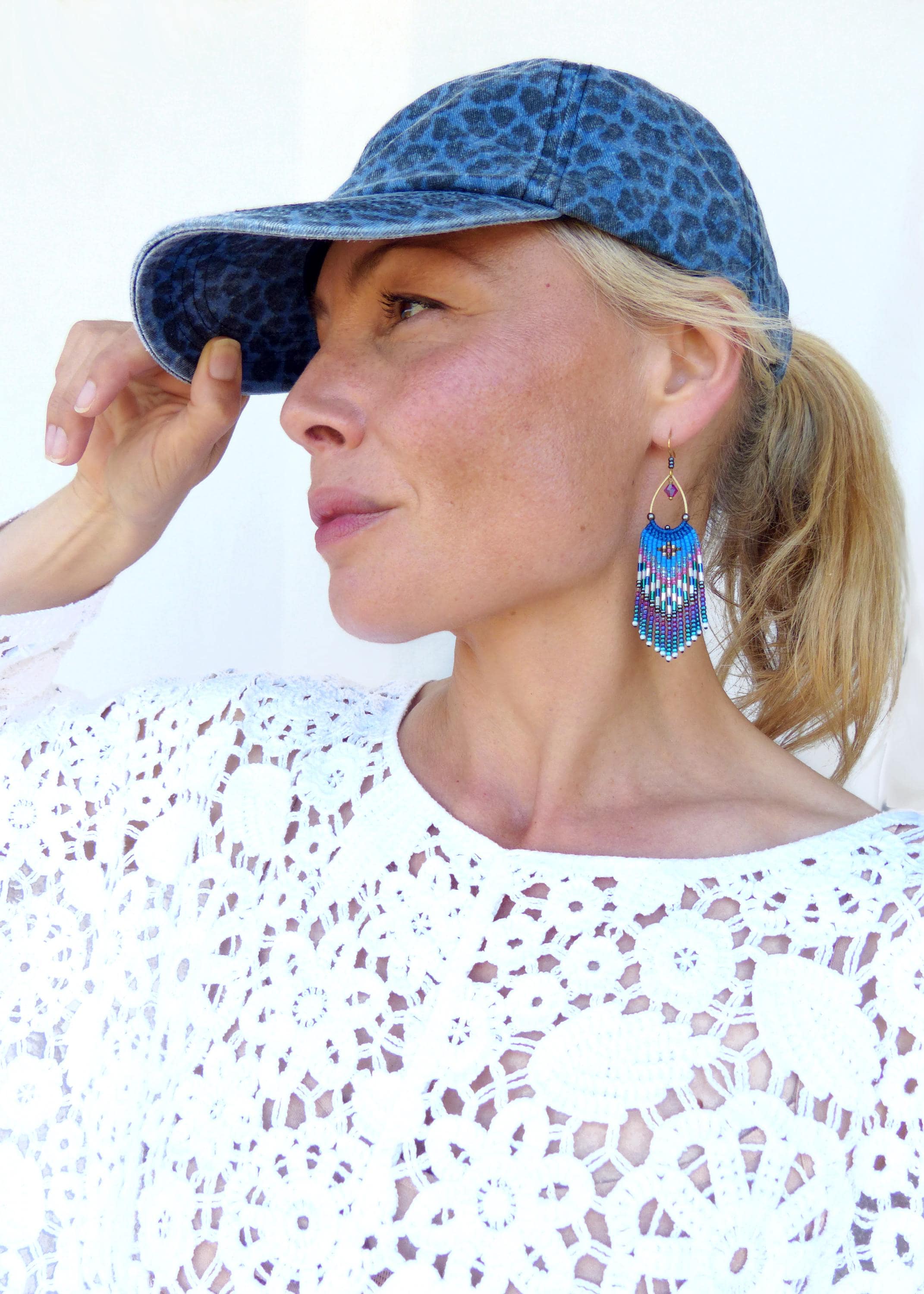 Peut inclure: Une personne porte une casquette de baseball en jean avec un imprimé léopard bleu. La personne porte également un haut en dentelle blanche et des boucles d'oreilles à franges bleues, violettes et blanches.