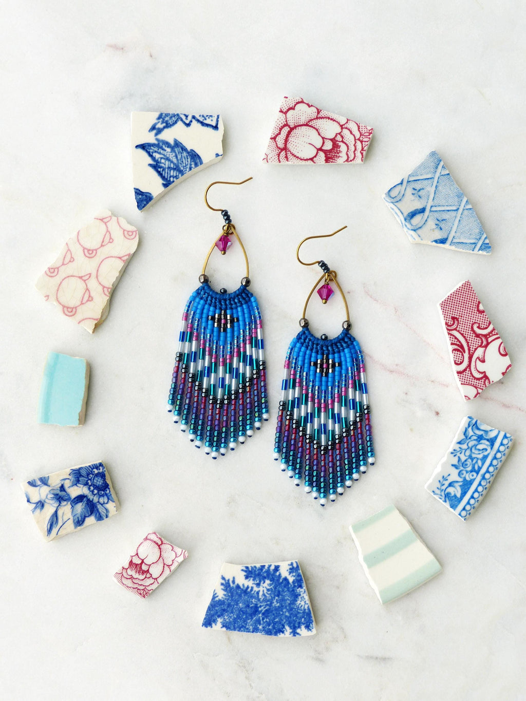 Peut inclure: Une paire de boucles d'oreilles en perles avec un motif de franges bleues, turquoise et violettes. Les boucles d'oreilles ont des cerceaux dorés et des accents de cristal rose. Elles sont entourées de fragments de céramique à motifs.