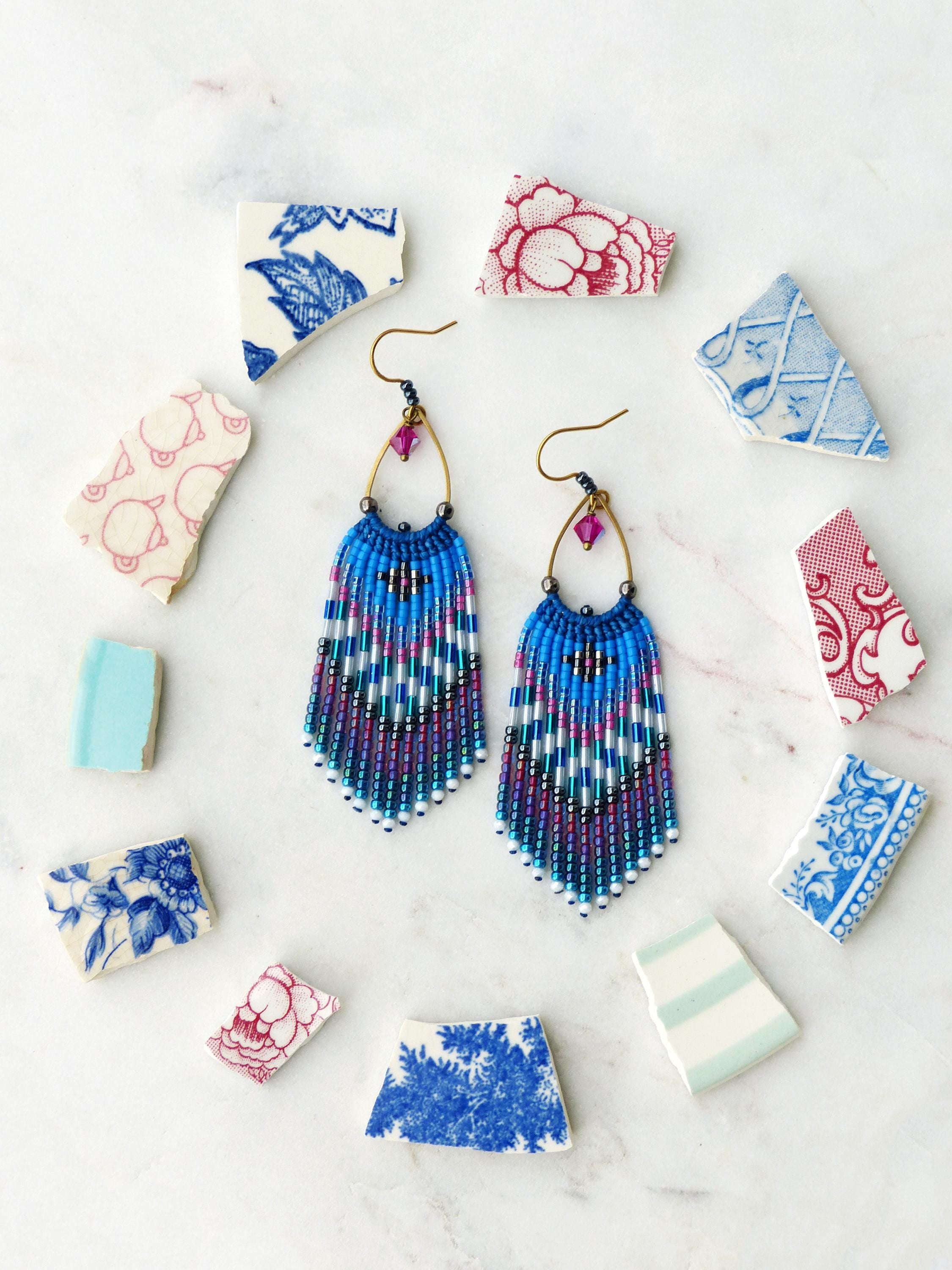 Peut inclure: Une paire de boucles d'oreilles en perles avec un motif de franges bleues, turquoise et violettes. Les boucles d'oreilles ont des cerceaux dorés et des accents de cristal rose. Elles sont entourées de fragments de céramique à motifs.