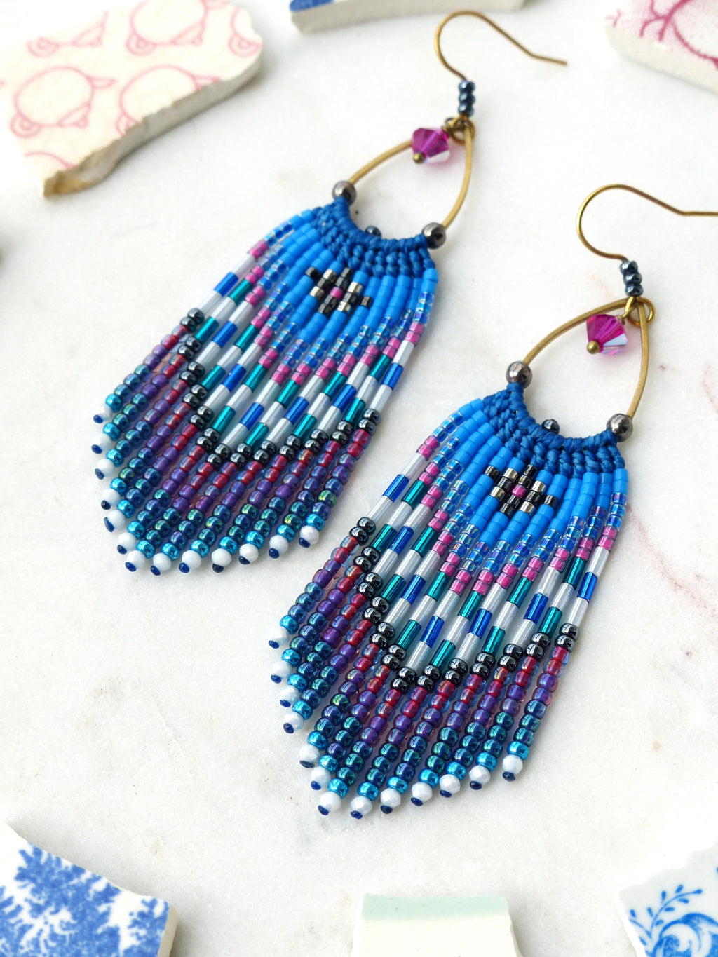 Peut inclure: Une paire de boucles d'oreilles à franges perlées dans les tons de bleu, violet, rose et argent. Les boucles d'oreilles présentent un motif géométrique et sont suspendues à des cerceaux dorés avec de petits accents roses.