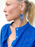 Peut inclure: Boucles d'oreilles en perles avec un anneau doré et une frange de perles bleues, blanches, violettes et turquoise. La personne porte une chemise bleue.