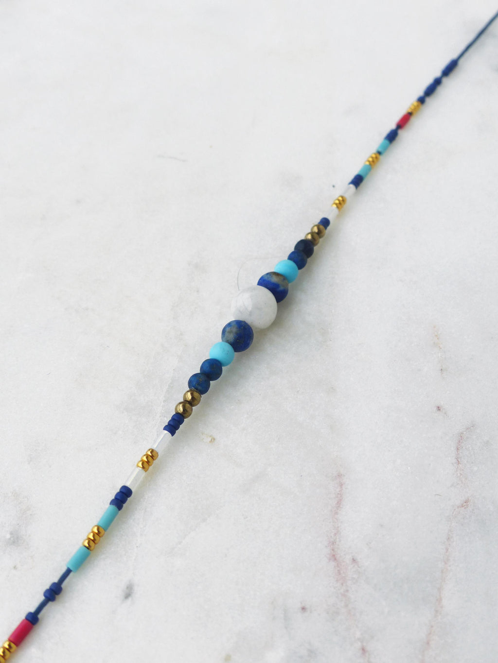 Peut inclure: Un collier de perles délicat avec un mélange de perles bleues, blanches et dorées. Le collier présente un groupe central de perles plus grandes dans les tons de bleu et de blanc, avec de plus petites perles dorées et bleues le long de la longueur. Sur une surface en marbre blanc.