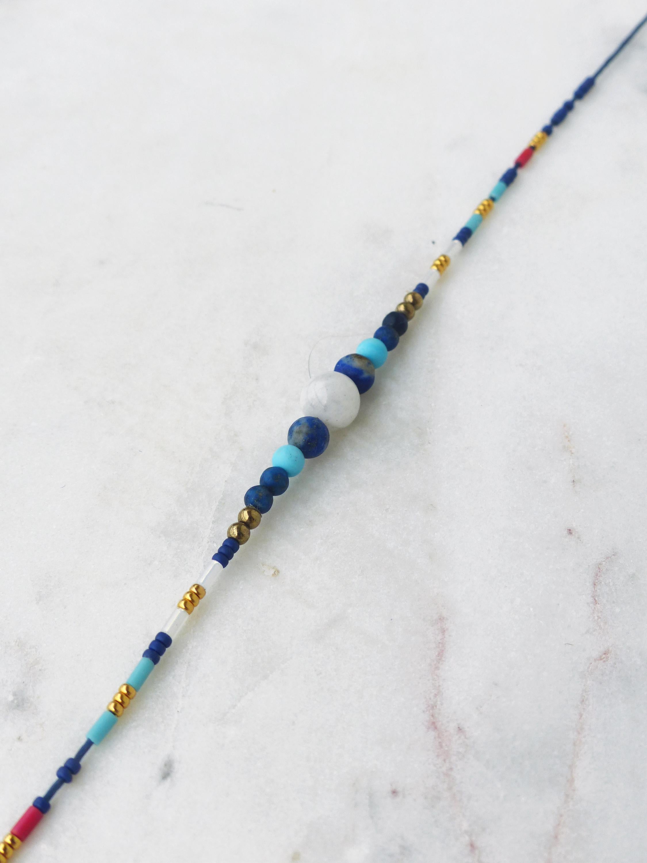 Peut inclure: Un collier de perles délicat avec un mélange de perles bleues, blanches et dorées. Le collier présente un groupe central de perles plus grandes dans les tons de bleu et de blanc, avec de plus petites perles dorées et bleues le long de la longueur. Sur une surface en marbre blanc.