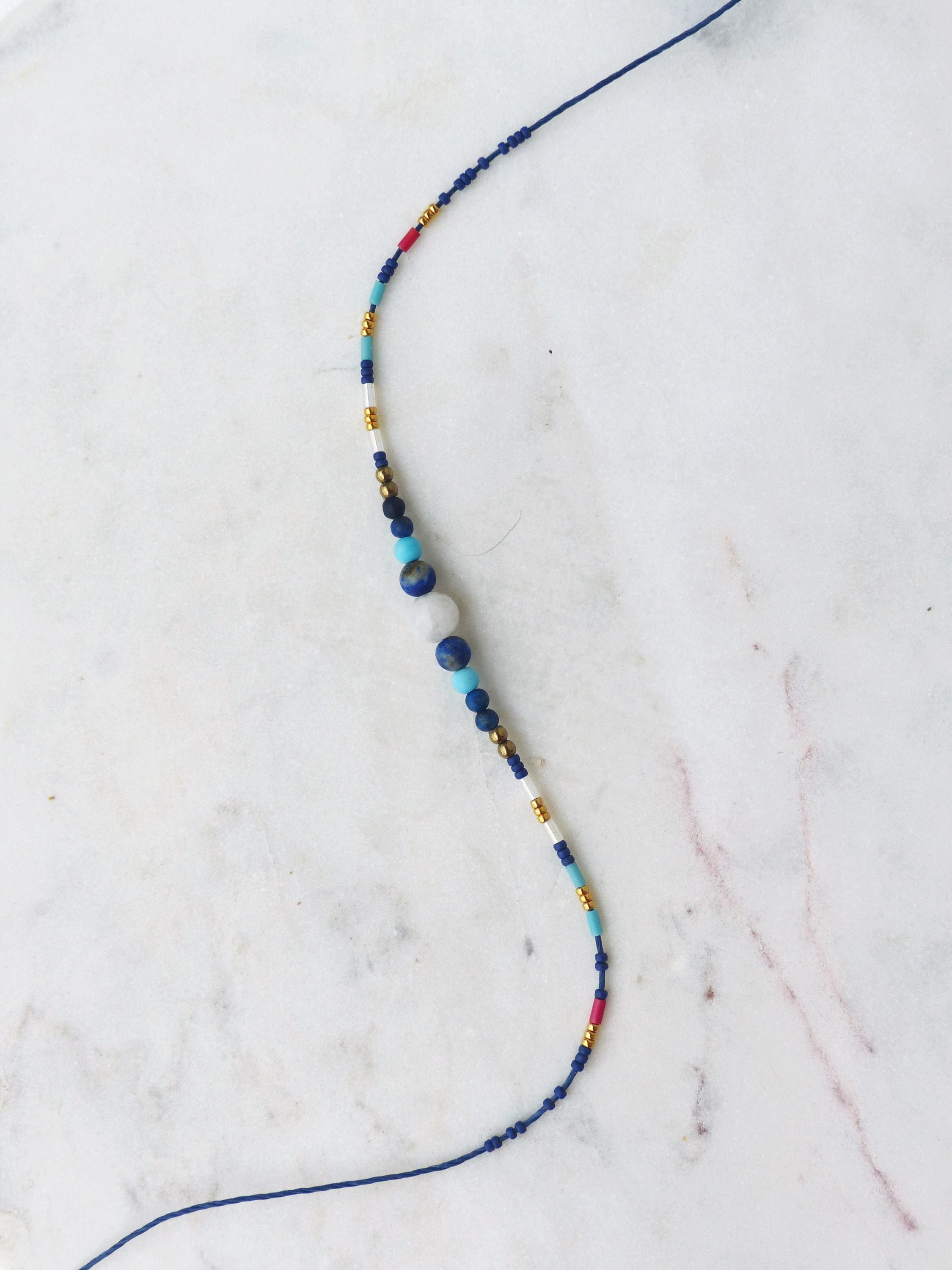 Peut inclure: Un collier délicat avec un cordon bleu foncé orné de diverses perles. Les perles comprennent de l'or, du rouge, du blanc, du turquoise et des nuances de bleu, avec une pierre blanche plus grande au centre. Le collier est de style minimaliste.