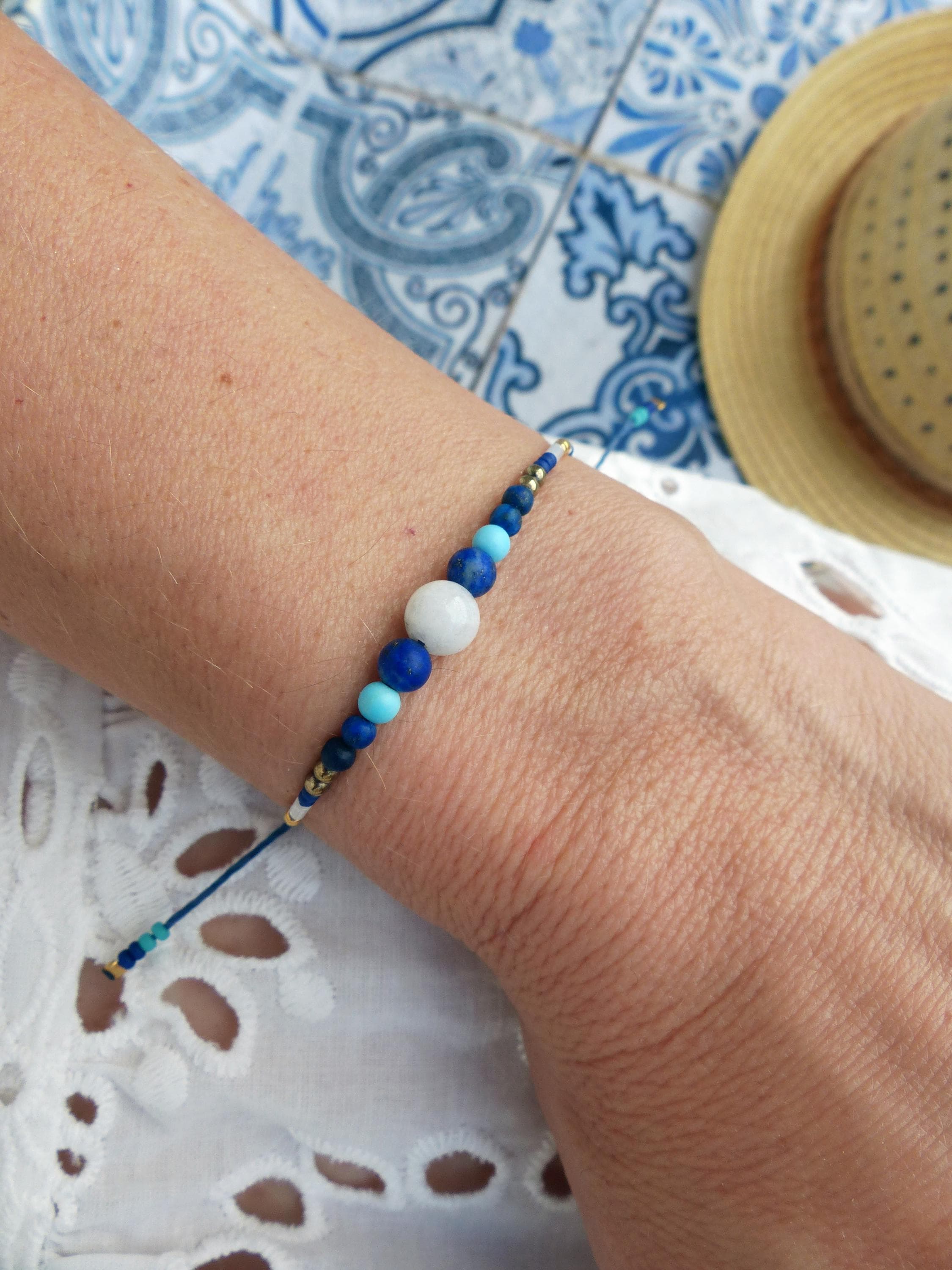 Peut inclure: Un bracelet de perles avec une grosse perle blanche, diverses perles bleues et des accents dorés. Le bracelet est monté sur un cordon bleu foncé. Le bracelet est sur le poignet d'une personne.