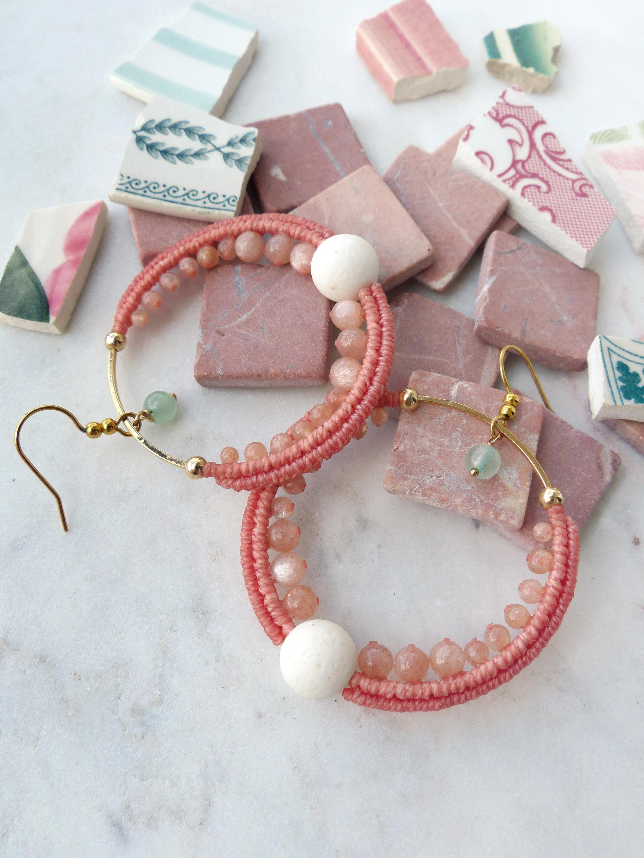 Peut inclure: Boucles d'oreilles créoles faites à la main. Elles présentent un design tissé de couleur corail, agrémenté de petites perles rondes pêche et d'une grosse perle blanche. Les boucles ont des accents dorés et une petite perle verte. Fond avec des carreaux roses et des fragments de céramique.