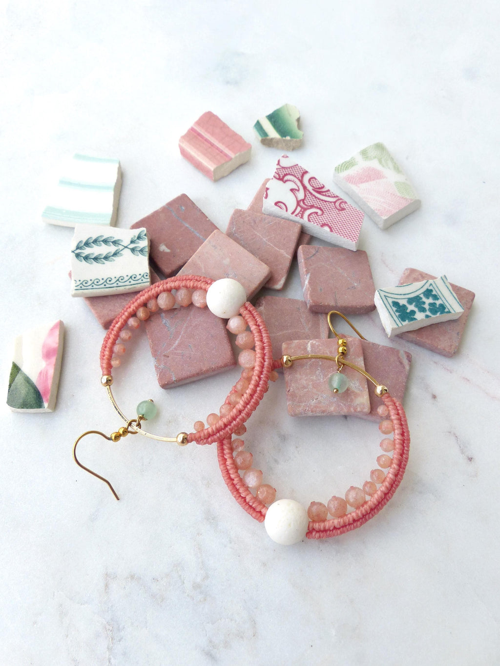 Peut inclure: Une paire de boucles d'oreilles créoles corail avec des crochets et des accents dorés. Les boucles d'oreilles sont ornées de petites perles et d'une grosse perle blanche. Elles sont présentées avec des carreaux roses et des morceaux de céramique décoratifs.