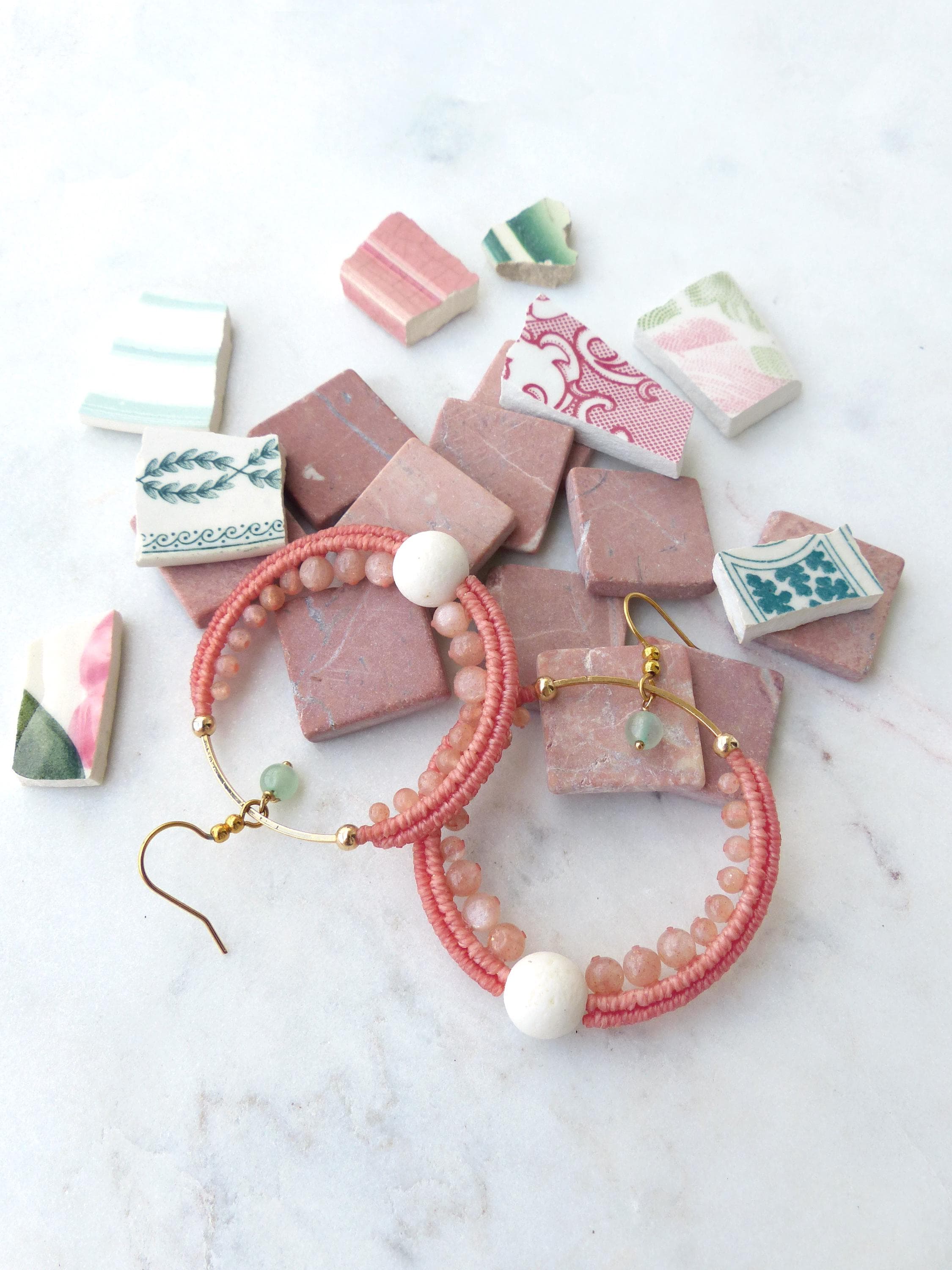 Peut inclure: Une paire de boucles d'oreilles créoles corail avec des crochets et des accents dorés. Les boucles d'oreilles sont ornées de petites perles et d'une grosse perle blanche. Elles sont présentées avec des carreaux roses et des morceaux de céramique décoratifs.