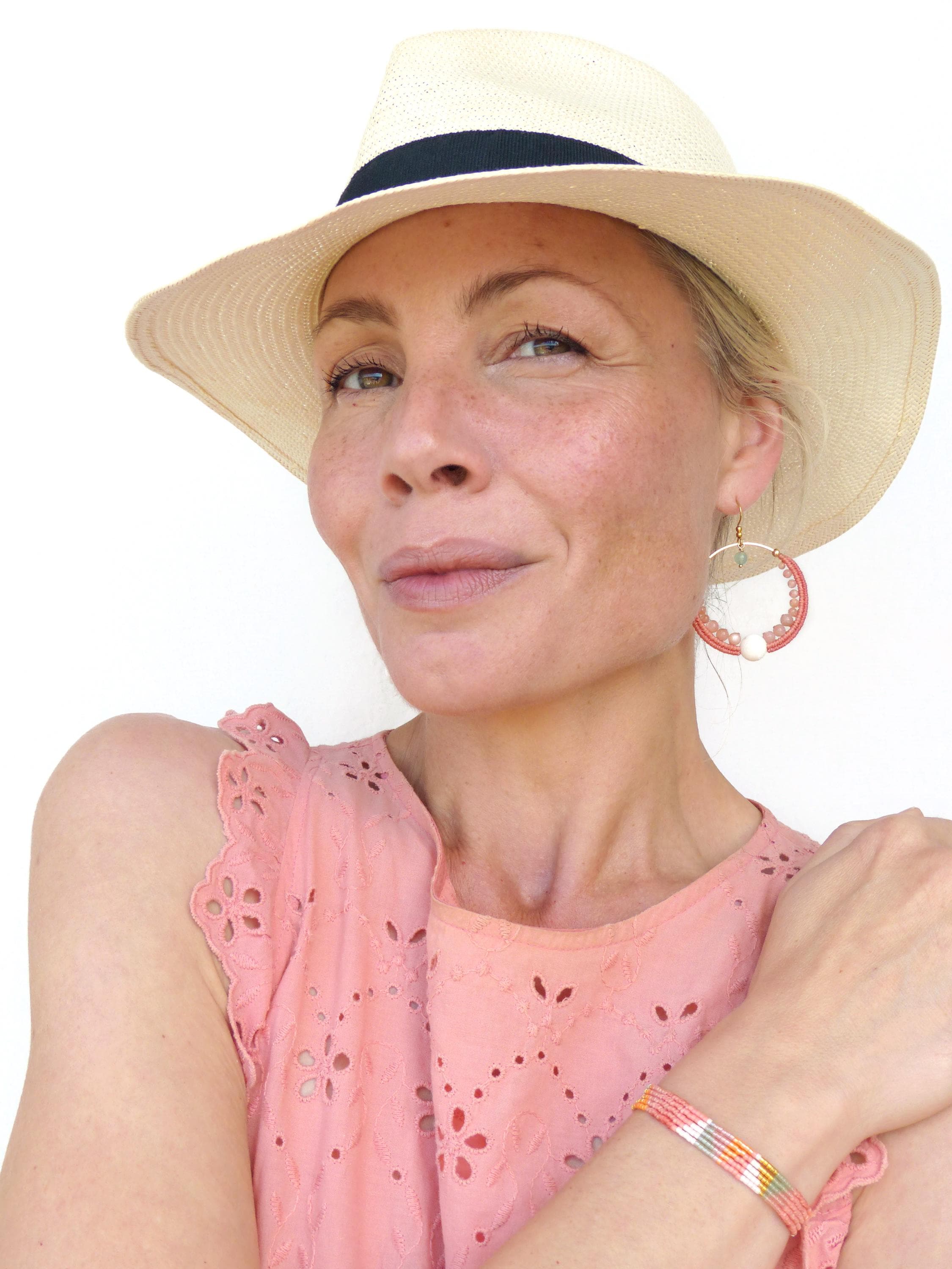 Peut inclure: Une femme porte un chapeau de paille crème avec une bande noire, un haut rose avec des découpes florales et des boucles d'oreilles créoles. Un bracelet coloré orne le poignet. Les boucles d'oreilles ont un design circulaire avec des accents rouges et blancs.