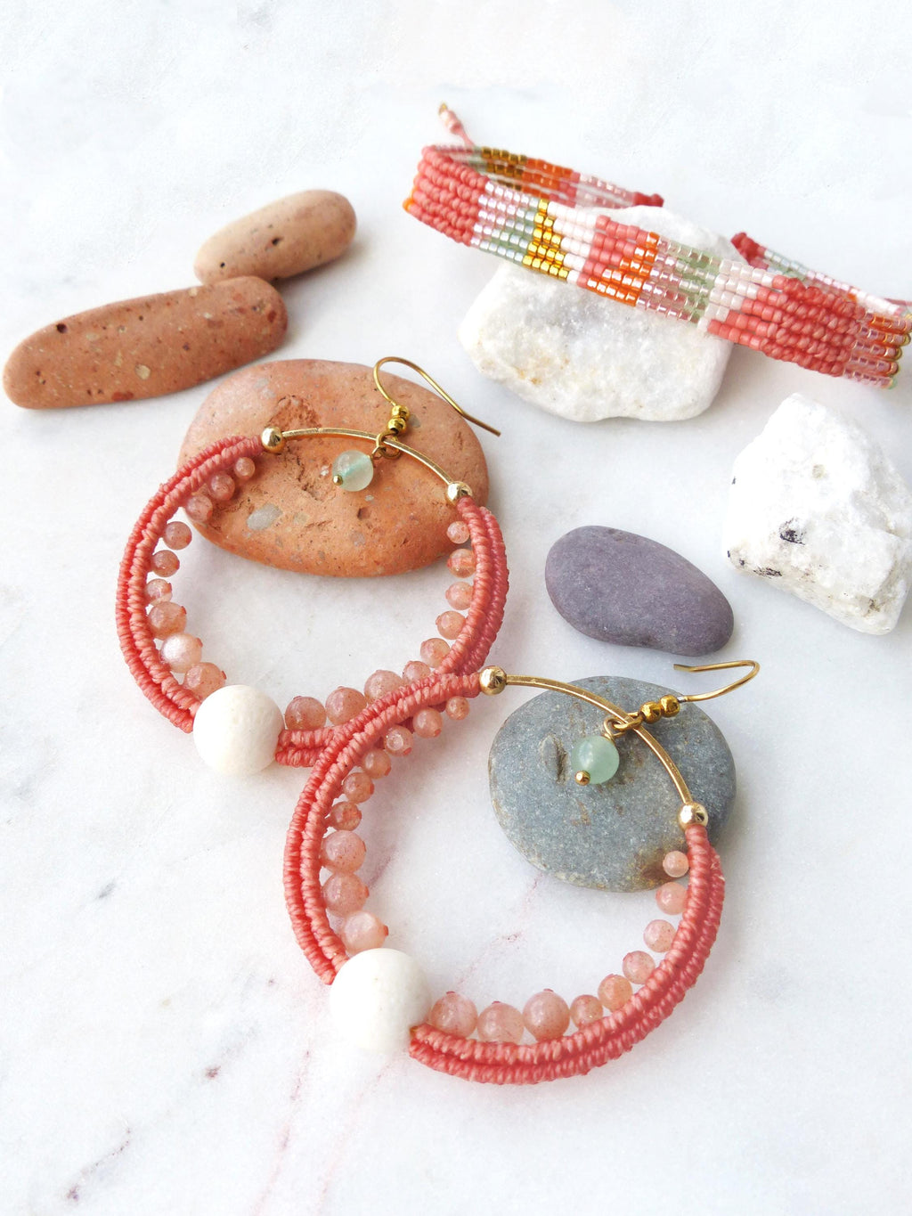 Peut inclure: Boucles d'oreilles créoles corail avec crochets dorés, ornées de petites perles et d'une grosse perle blanche. Un bracelet tissé assorti et des pierres décoratives complètent l'image. Les bijoux sont présentés sur une surface blanche.