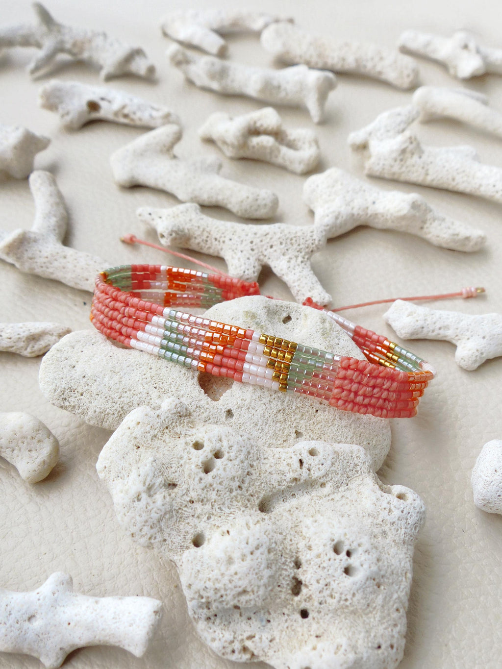 Peut inclure: Un bracelet de perles tissées aux couleurs corail, blanc, or et vert. Le bracelet est posé sur une pierre blanche poreuse, entouré de morceaux de corail blanc. Le bracelet est réglable avec une ficelle rose.