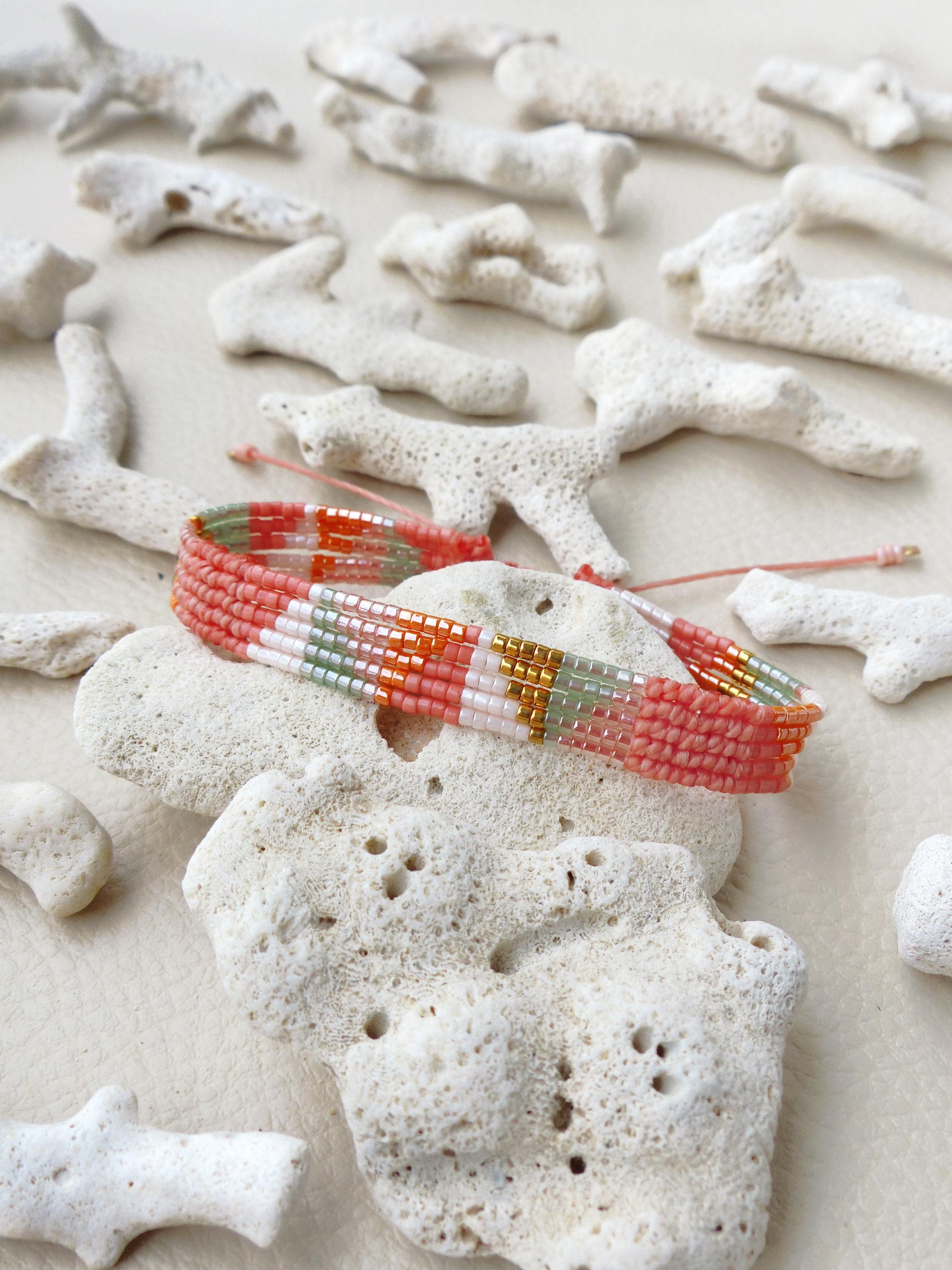 Peut inclure: Un bracelet de perles tissées aux couleurs corail, blanc, or et vert. Le bracelet est posé sur une pierre blanche poreuse, entouré de morceaux de corail blanc. Le bracelet est réglable avec une ficelle rose.