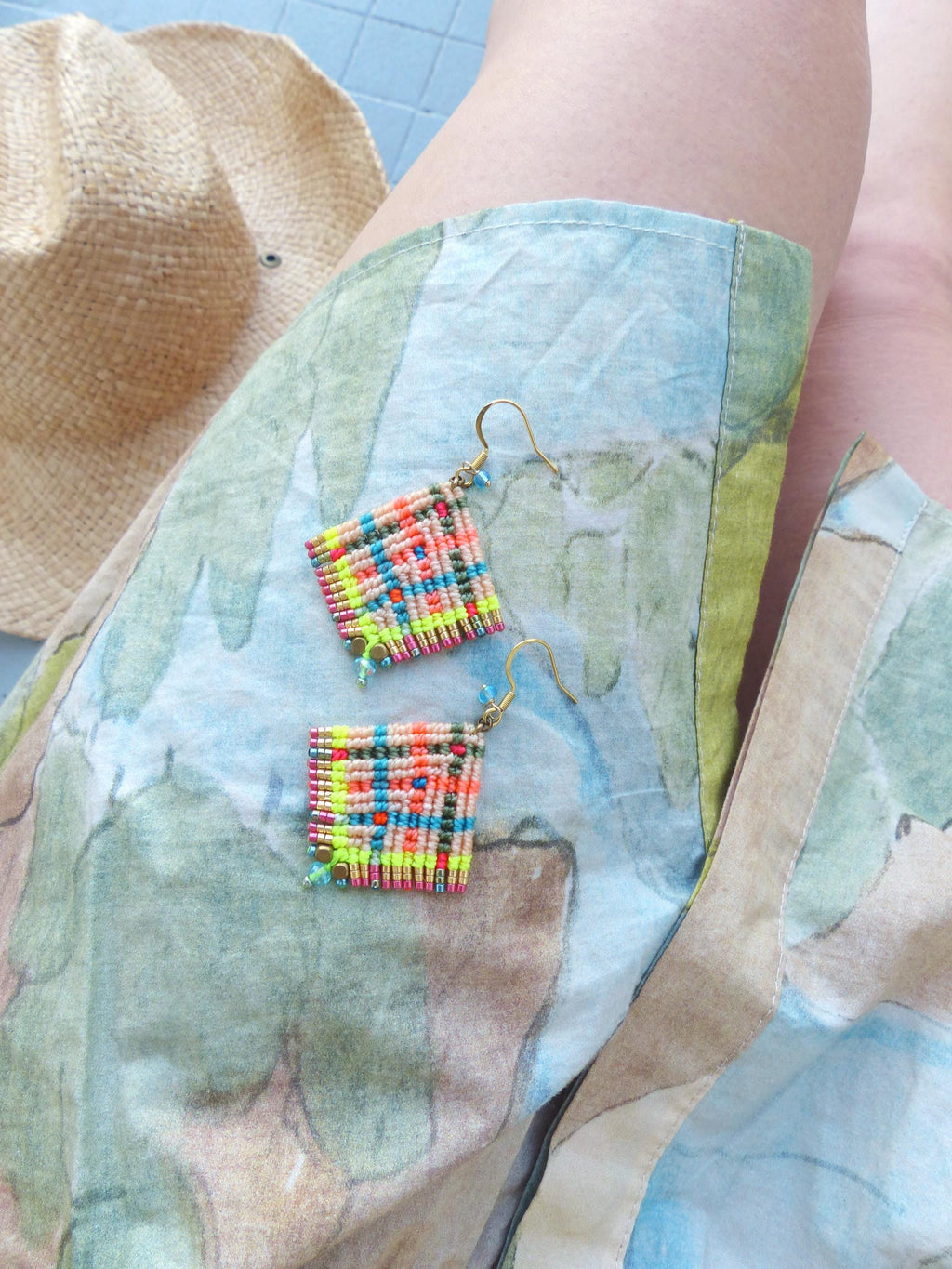 Peut inclure: Une paire de boucles d'oreilles carrées en macramé avec des crochets dorés. Les boucles d'oreilles présentent un motif tissé coloré avec des fils jaune fluo, orange, rose et turquoise. Un chapeau de paille est en arrière-plan.