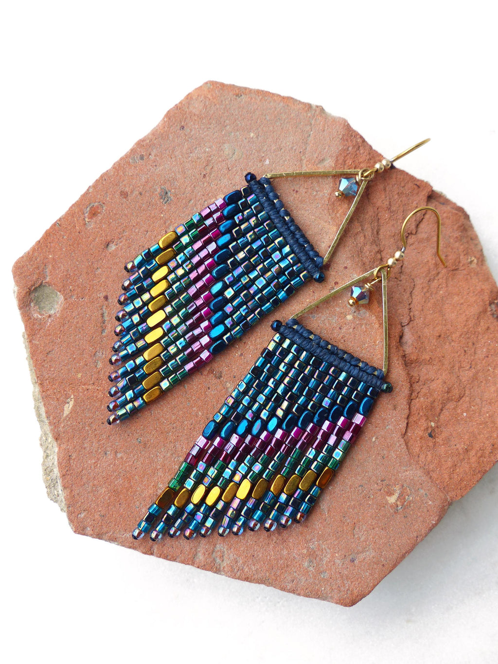 Peut inclure: Une paire de boucles d'oreilles perlées faites à la main avec un design géométrique. Les boucles d'oreilles présentent un cadre triangulaire doré et une frange de perles colorées dans les tons de bleu, violet, vert et or. Elles sont posées sur une surface texturée brun-rouge.