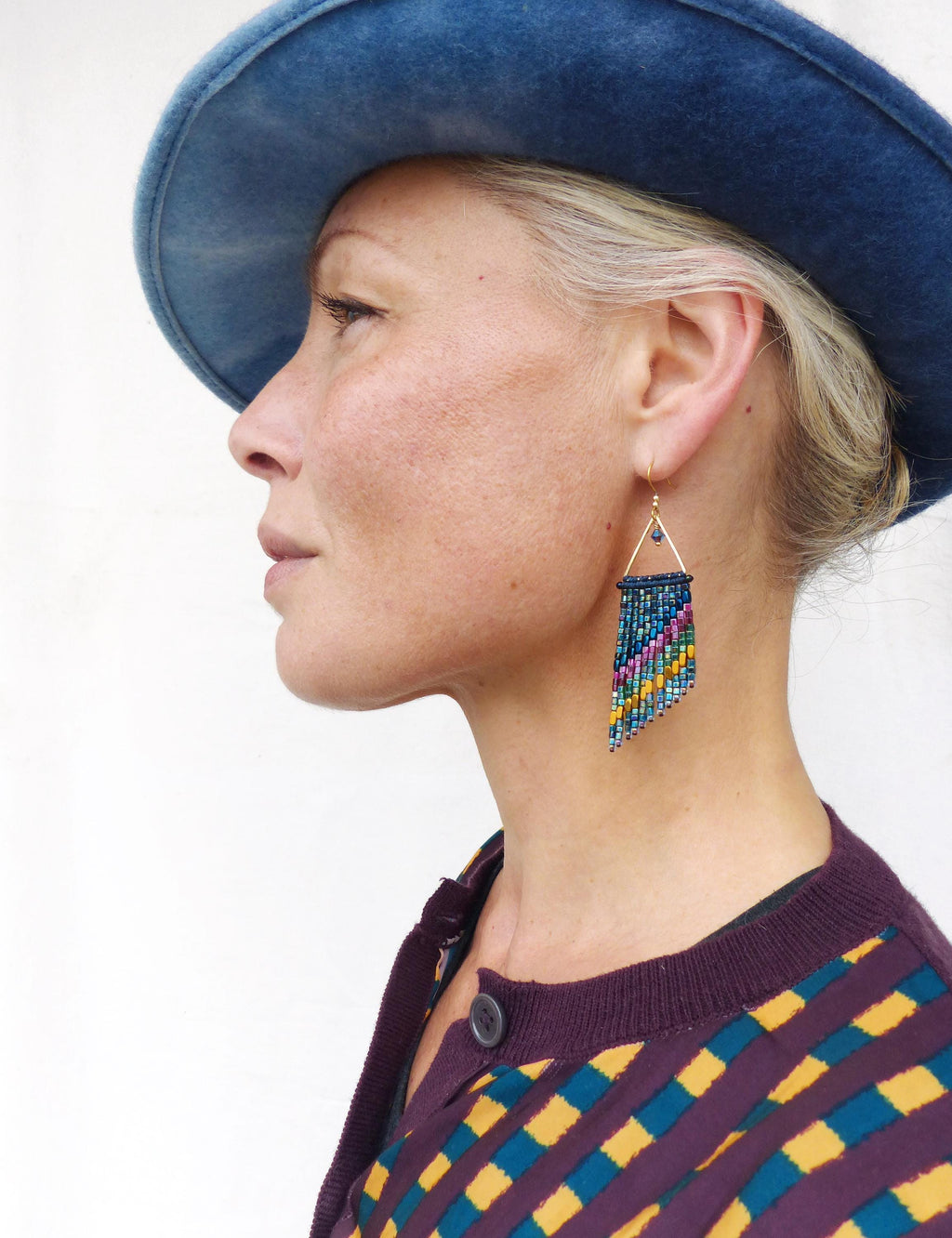 Peut inclure: Profil d'une personne portant un chapeau bleu et un cardigan violet à motifs géométriques. La personne porte des boucles d'oreilles en perles avec un dessus triangulaire doré et une frange de perles bleues, roses et jaunes.
