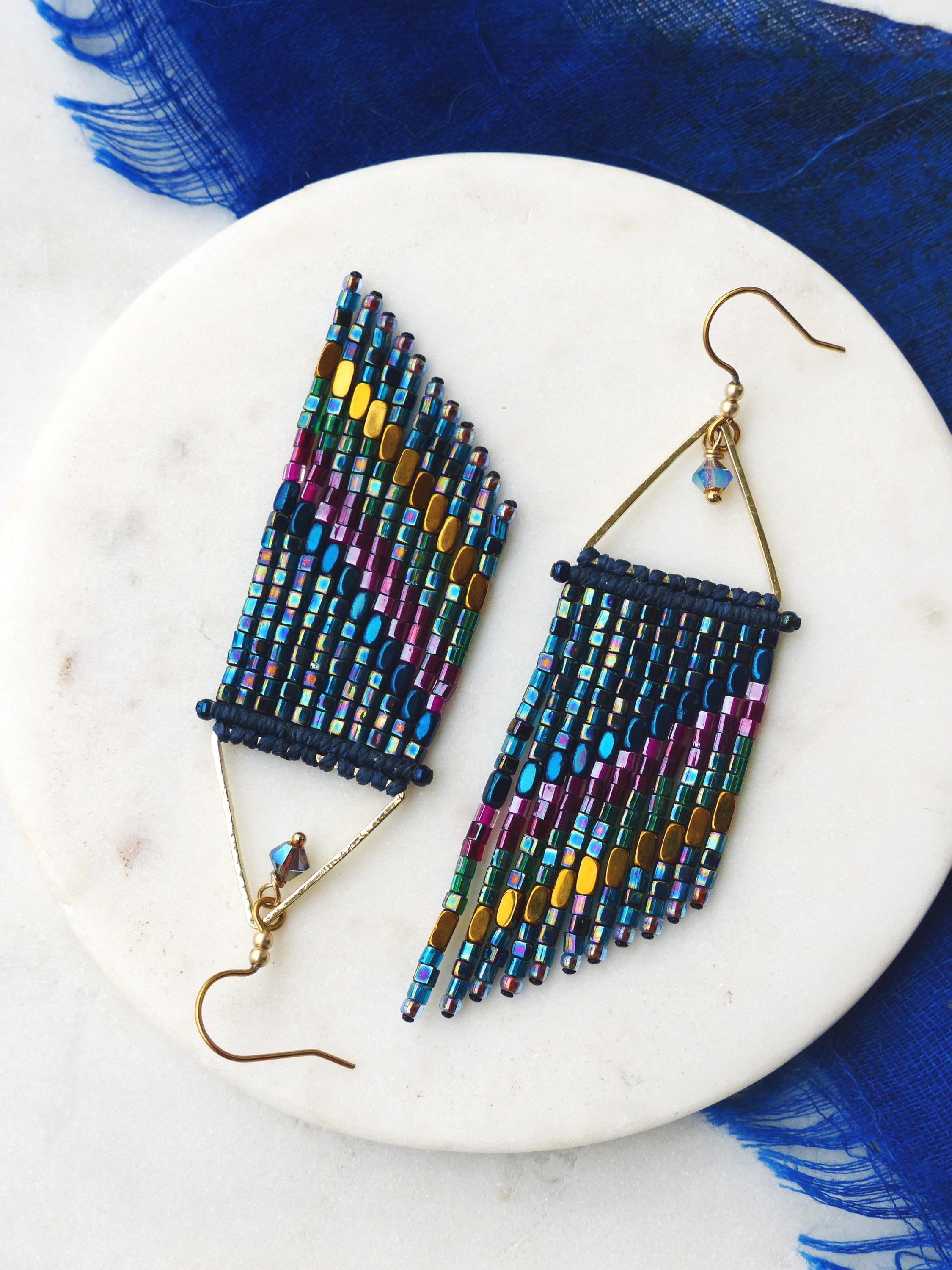 Peut inclure: Une paire de boucles d'oreilles perlées faites à la main. Les boucles d'oreilles présentent un motif de franges avec un mélange de perles bleues, violettes, dorées et vertes. Elles ont des montures triangulaires et des crochets d'oreilles dorés. 
