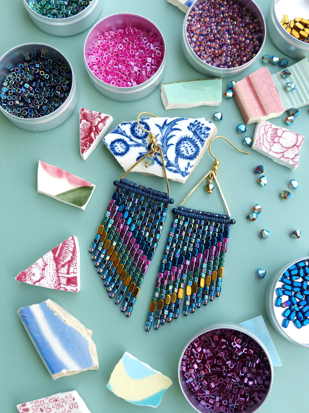 Peut inclure: Boucles d'oreilles à franges de perles avec des crochets dorés. Les boucles d'oreilles présentent un mélange de perles bleues, violettes et dorées. Autour, de petites boîtes remplies de perles colorées et des morceaux de carreaux de céramique cassés.