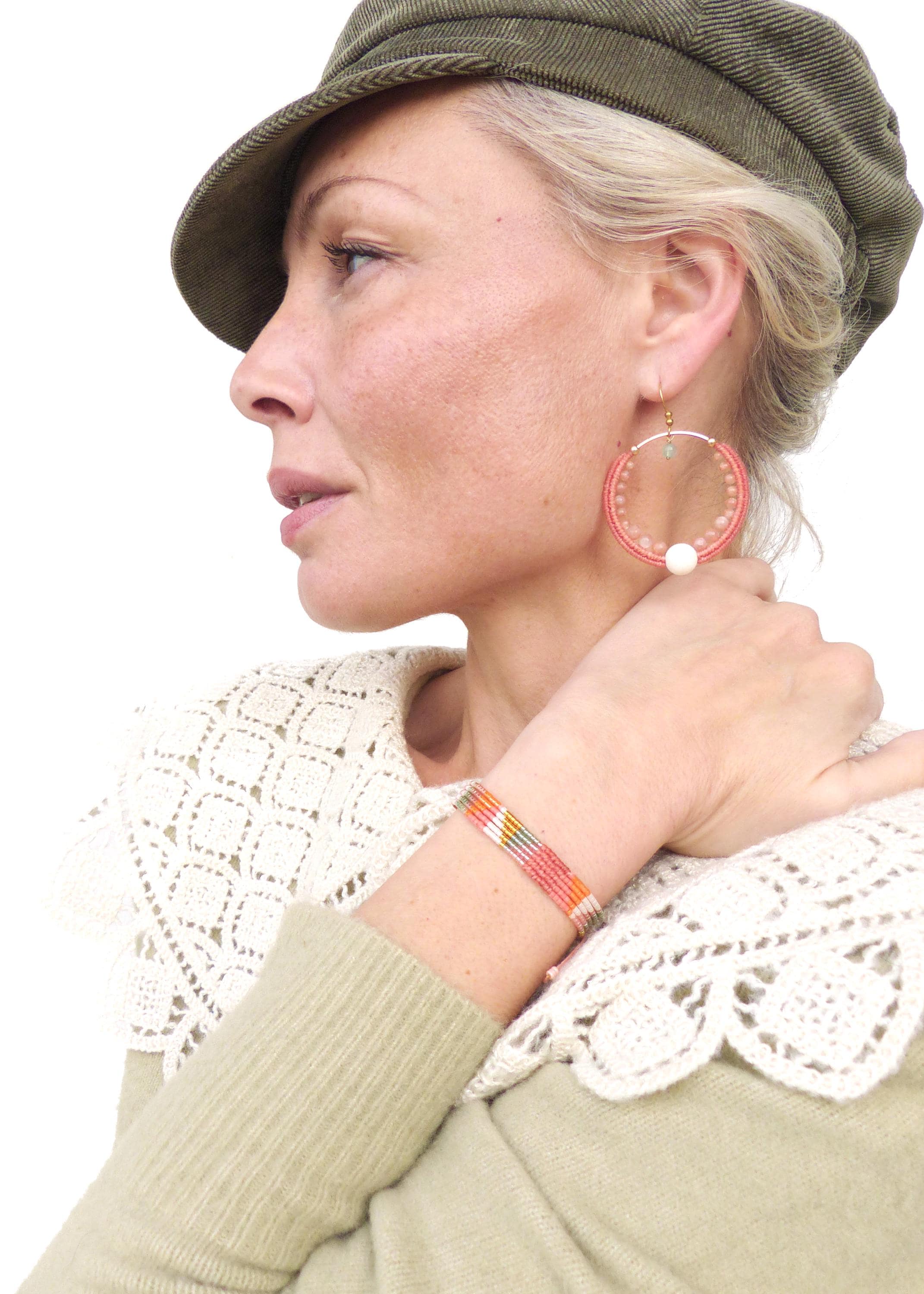 Peut inclure: Une femme porte une casquette verte, un pull crème et un col en dentelle blanc. Elle porte de grandes boucles d'oreilles créoles avec une perle et un bracelet de perles rayé orange, rose et blanc. Les bijoux sont faits à la main.