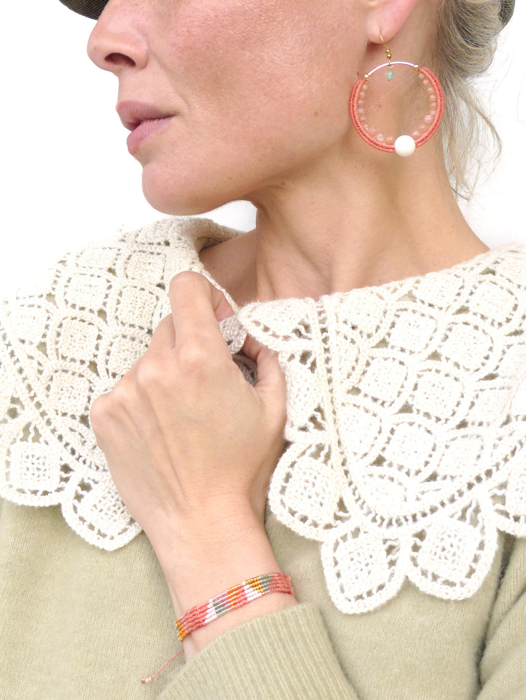 Peut inclure: Gros plan d'une personne portant un col en crochet crème et un bracelet avec des fils rouges, jaunes et blancs. La personne porte également de grandes boucles d'oreilles créoles avec des perles roses et une perle blanche.