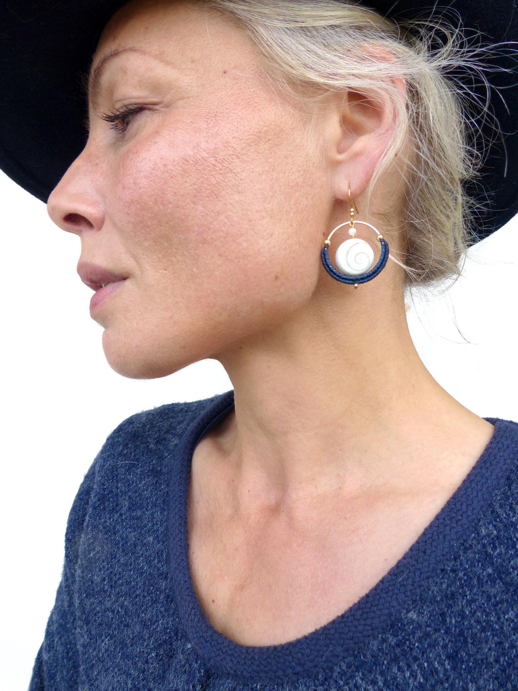 Peut inclure: Boucles d'oreilles dorées avec un motif en spirale blanc et des accents bleu marine. Les boucles d'oreilles sont ornées d'une petite perle dorée et sont portées par une personne vêtue d'un pull bleu.