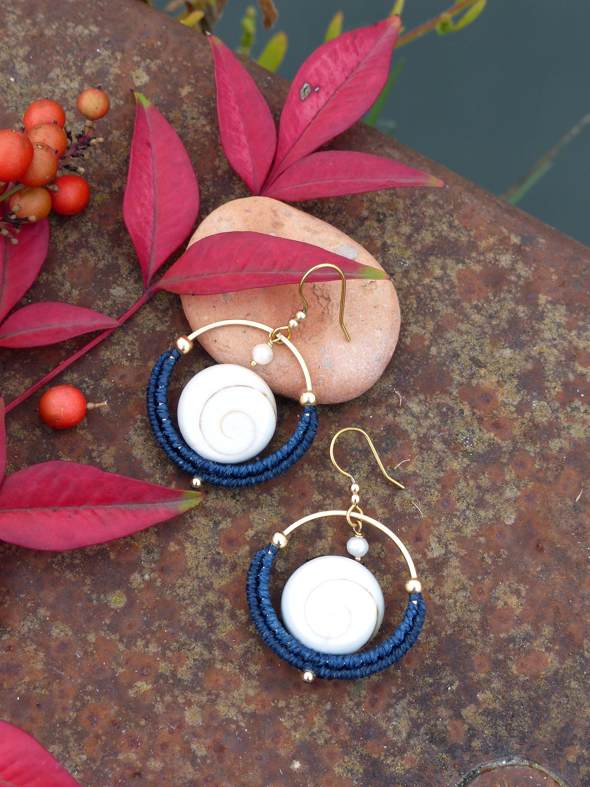 Peut inclure: Boucles d'oreilles artisanales avec un cerceau doré, chacune ornée d'un centre blanc en spirale entouré de fil bleu marine. Les boucles d'oreilles sont posées sur une surface brun rouille, avec des feuilles et des baies rouges en arrière-plan. Bijoux uniques.