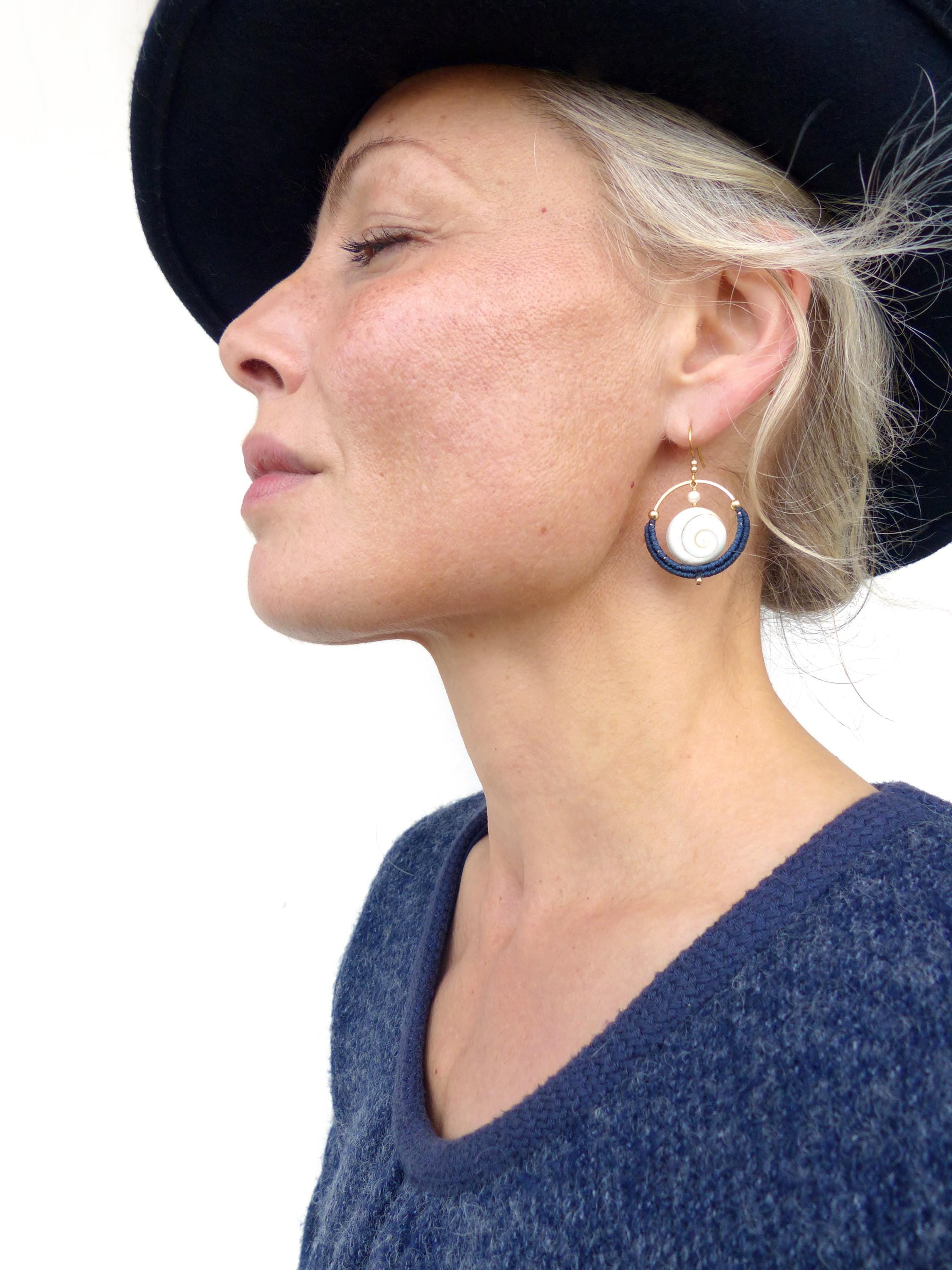Peut inclure: Boucles d'oreilles dorées avec un centre blanc en forme de coquillage et des détails bleu marine. Les boucles d'oreilles sont suspendues à des crochets dorés. La personne porte un pull bleu marine et un chapeau noir.