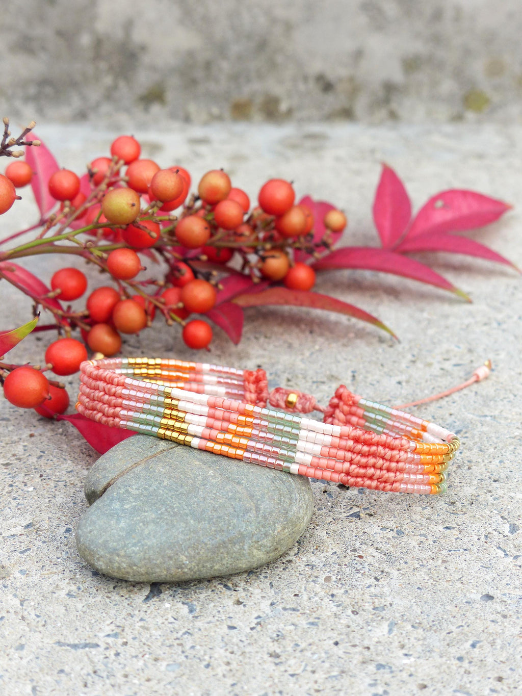 Peut inclure: Un bracelet tissé coloré avec une fermeture macramé, présentant un motif de fils roses, oranges, verts et dorés. Le bracelet repose sur une pierre grise, avec des baies et des feuilles rouges en arrière-plan.