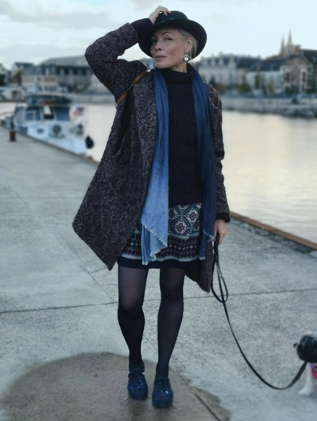 Peut inclure: Une personne porte un chapeau noir, un manteau foncé et une jupe à motifs sur un quai. Elle porte une écharpe bleue et des chaussures noires. Un petit chien en laisse est visible, avec des bateaux et des bâtiments en arrière-plan.