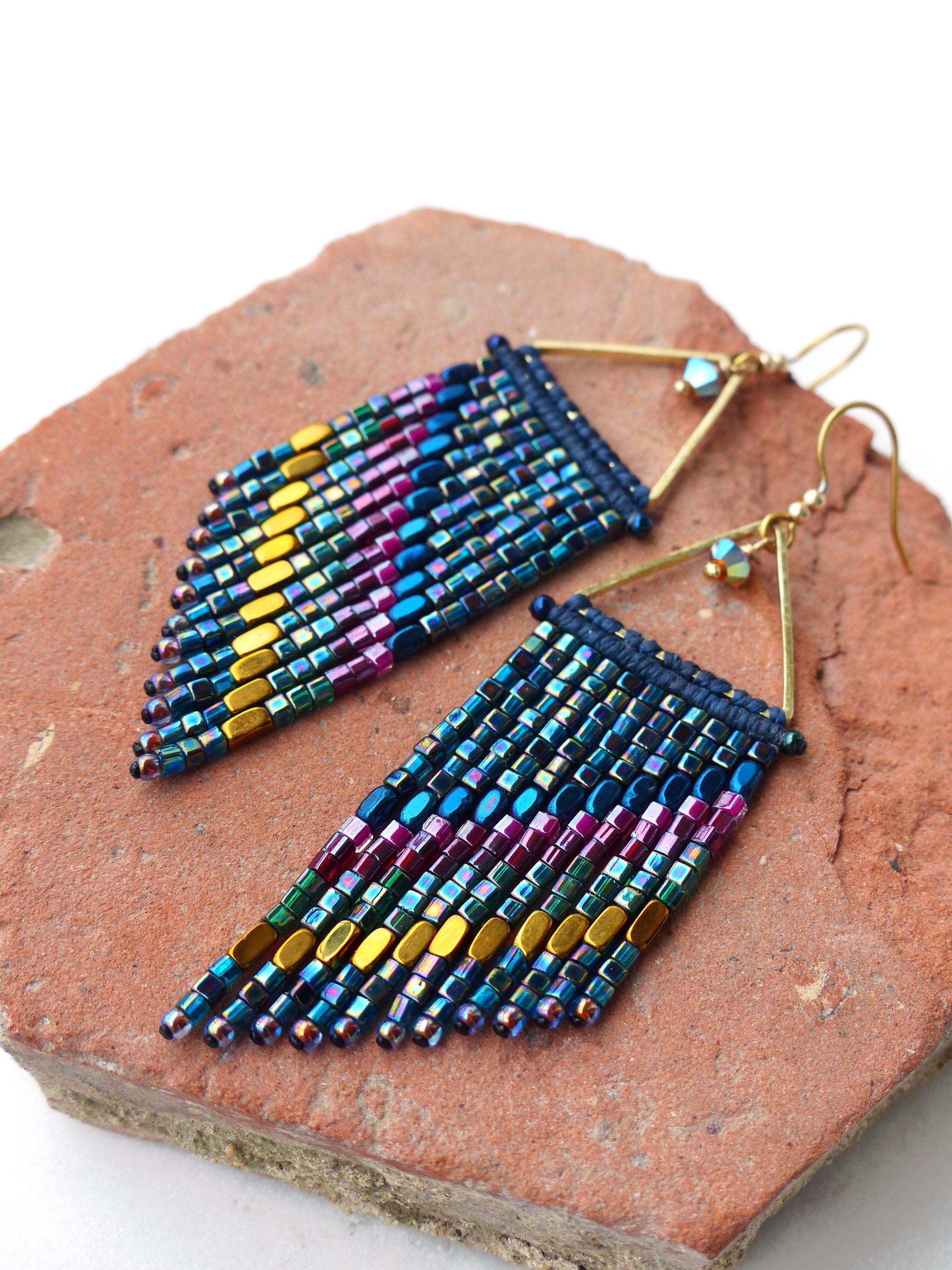 Peut inclure: Une paire de boucles d'oreilles perlées faites à la main avec un design à franges. Les boucles d'oreilles présentent un mélange de perles irisées dans les tons de bleu, violet, vert et or. Elles sont suspendues à des montures triangulaires dorées et sont présentées sur une surface brun-rouge.