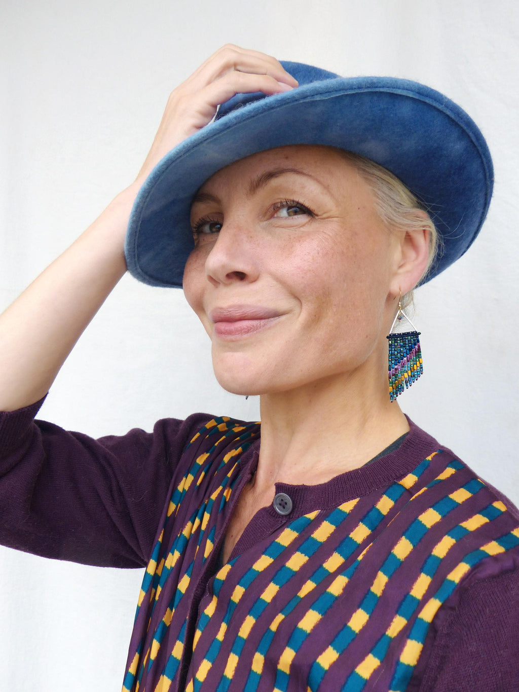 Peut inclure: Une personne portant un chapeau bleu et un cardigan violet avec un motif géométrique jaune et turquoise. Elle porte également des boucles d'oreilles en perles avec un cadre triangulaire argenté et des perles pendantes bleues, violettes et jaunes.