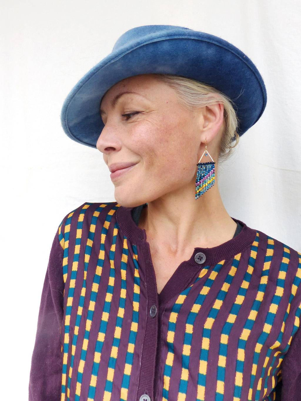 Peut inclure: Une femme porte un chapeau bleu et un cardigan violet à motif géométrique jaune et turquoise. Elle porte également des boucles d'oreilles en perles avec un haut triangulaire et une frange colorée.