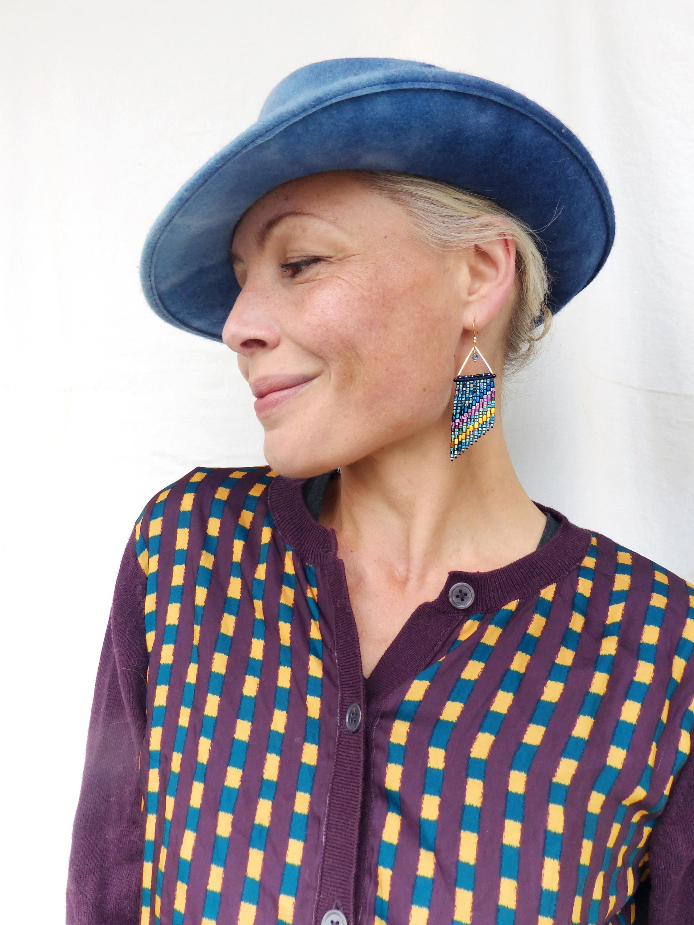 Peut inclure: Une femme porte un chapeau bleu et un cardigan violet à motif géométrique jaune et turquoise. Elle porte également des boucles d'oreilles en perles avec un haut triangulaire et une frange colorée.