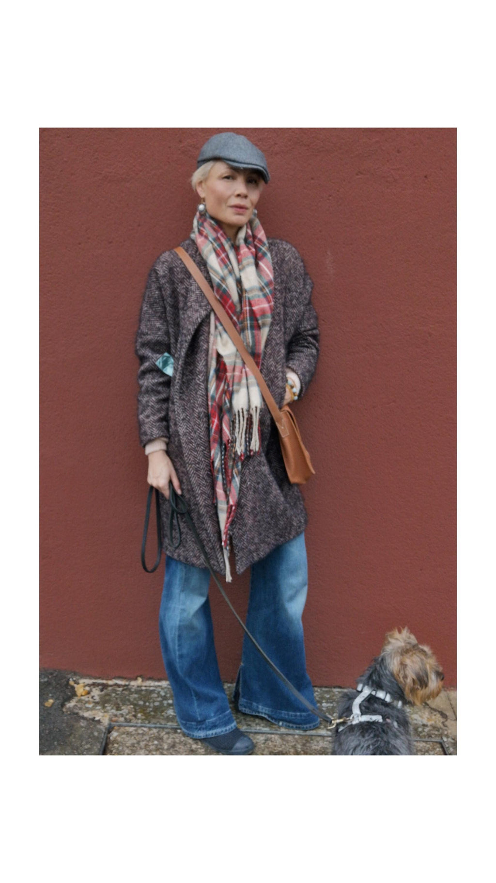 Peut inclure: Une femme porte un manteau en tweed gris, un jean bleu et une écharpe à carreaux. Elle porte également une casquette grise et un sac bandoulière en cuir marron. Un petit chien est en laisse.