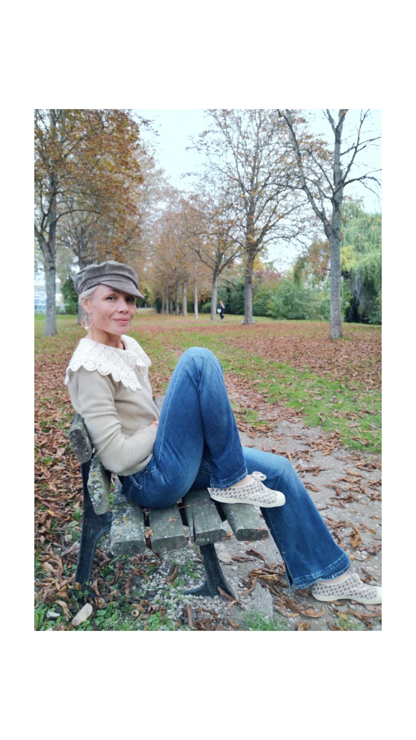 Peut inclure: Une personne vêtue d'un pull beige avec un col en dentelle, d'un jean bleu et de chaussures à motifs est assise sur un banc de parc. Elle porte une casquette grise. L'arrière-plan présente des arbres aux feuilles d'automne et un sentier.