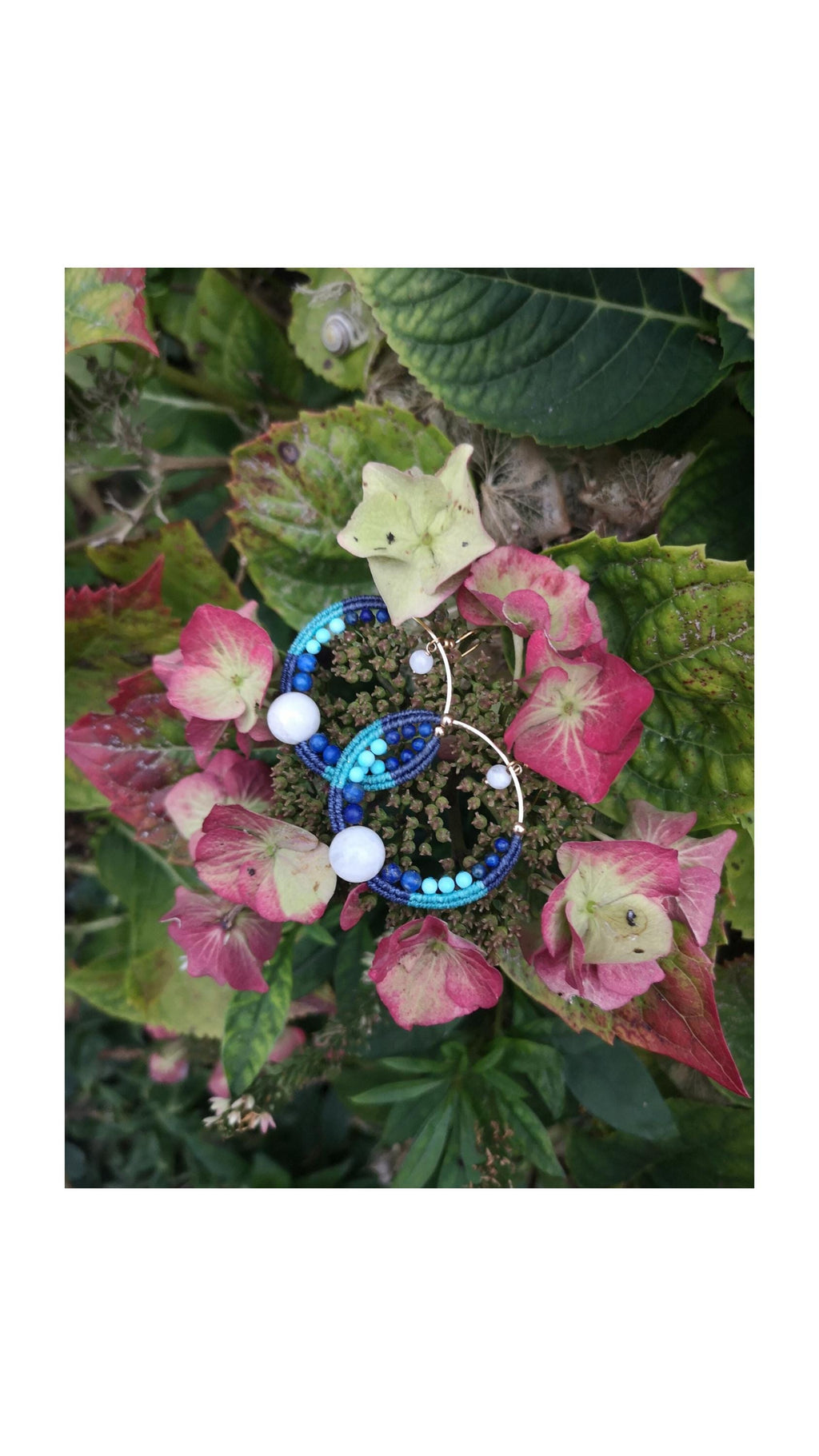 Peut inclure: Boucles d'oreilles créoles faites à la main avec un design macramé. Les boucles d'oreilles présentent des perles bleues et blanches, avec une grosse perle blanche en bas. Elles sont posées sur un fond de feuillage rose et vert.