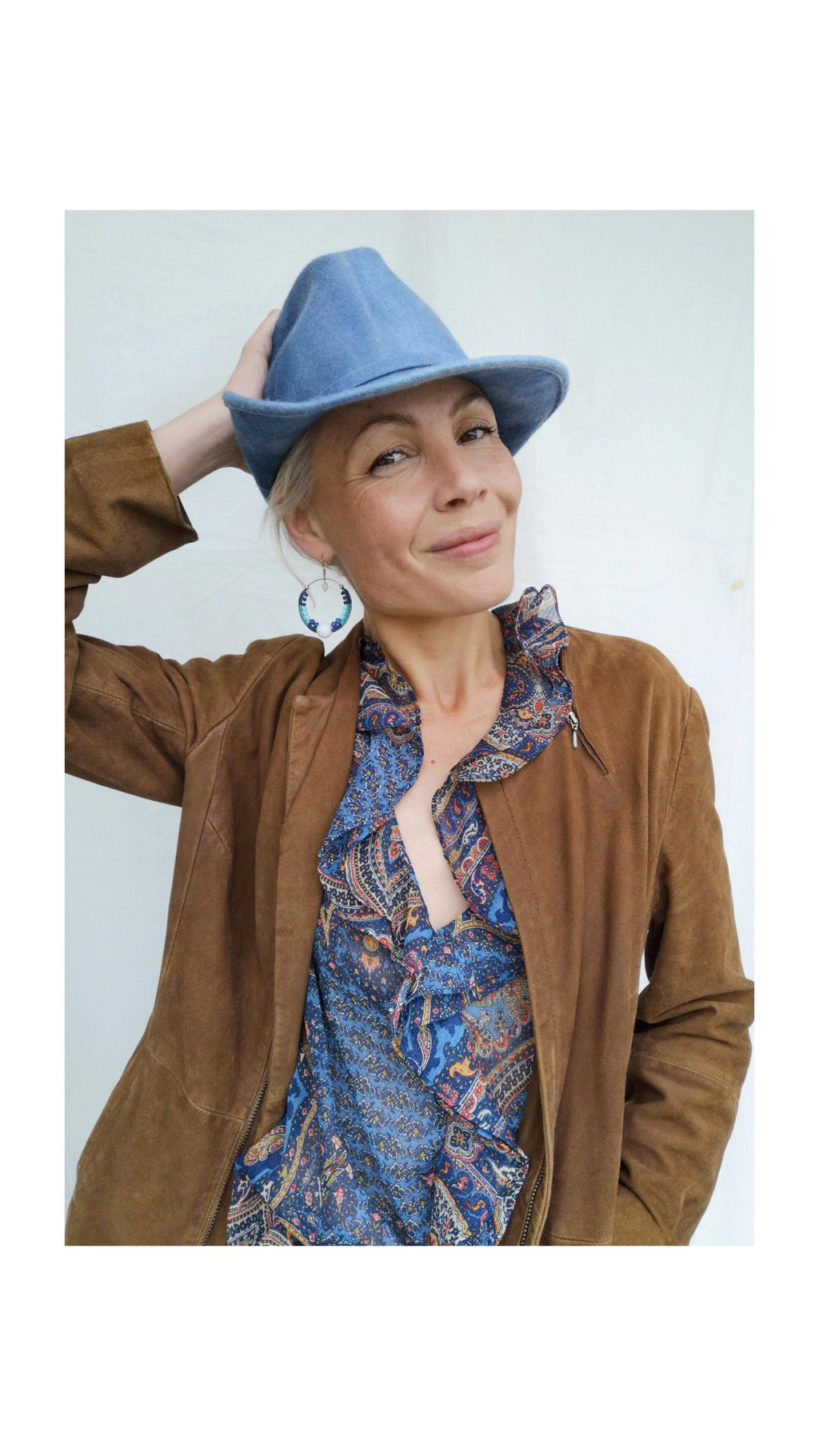 Peut inclure: Une femme portant un chapeau bleu clair, une veste en daim marron et un chemisier à imprimé cachemire bleu. La veste a une fermeture éclair. Le chemisier a une encolure à volants. La femme porte également des boucles d'oreilles créoles.