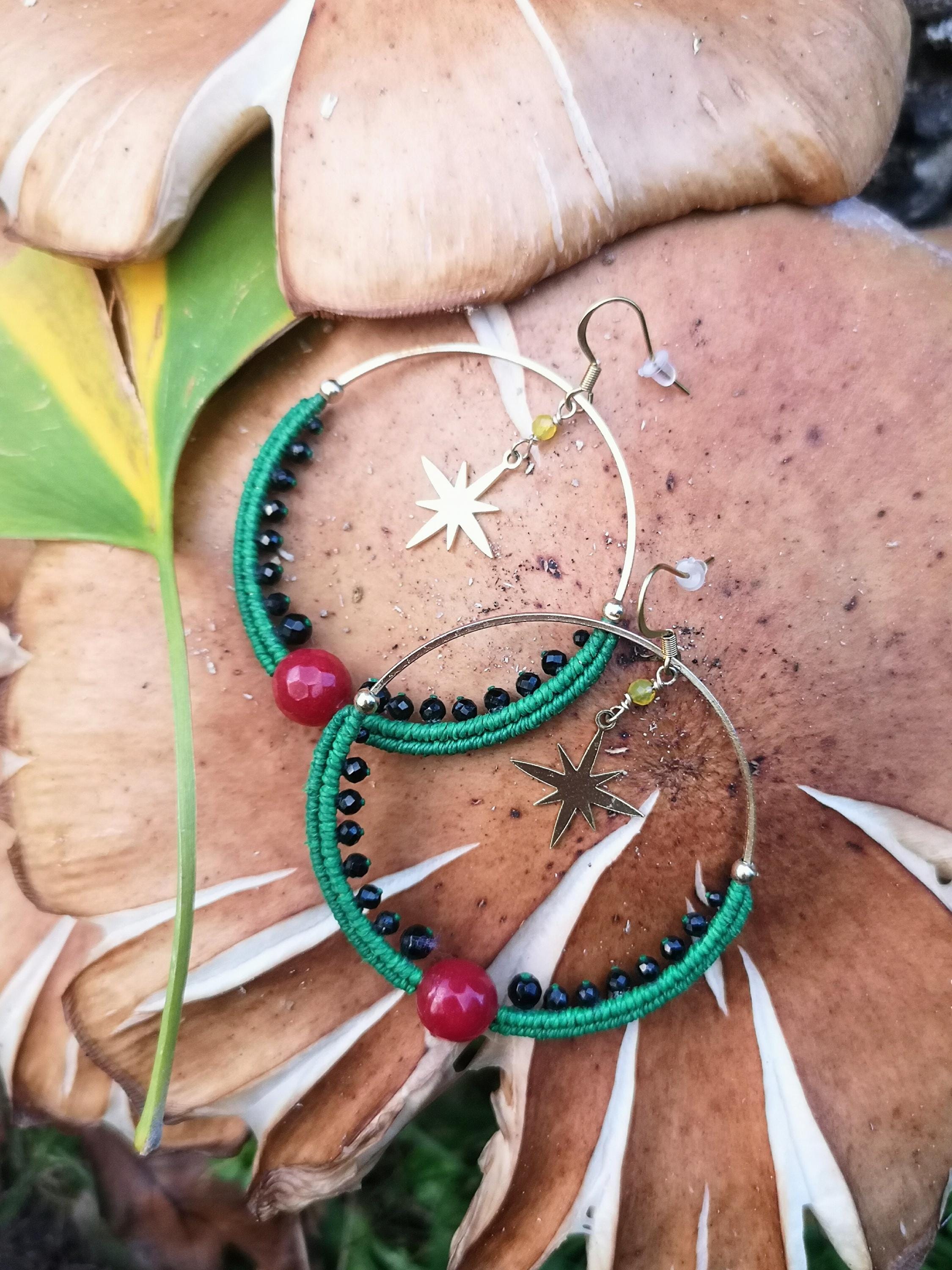 Peut inclure: Boucles d'oreilles créoles dorées avec macramé vert, perles noires et perles rouges. Chaque boucle d'oreille est ornée d'un charm en forme d'étoile dorée. Les boucles d'oreilles sont présentées sur un champignon.
