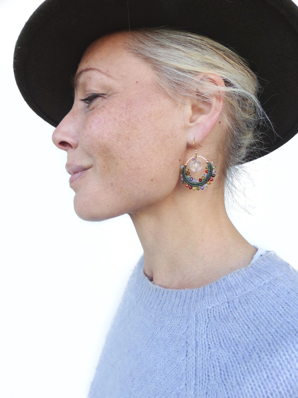 Peut inclure: Boucles d'oreilles créoles dorées avec un cristal transparent et des perles colorées. Les boucles d'oreilles sont attachées à un crochet doré. La femme porte un pull bleu clair et un chapeau marron foncé.