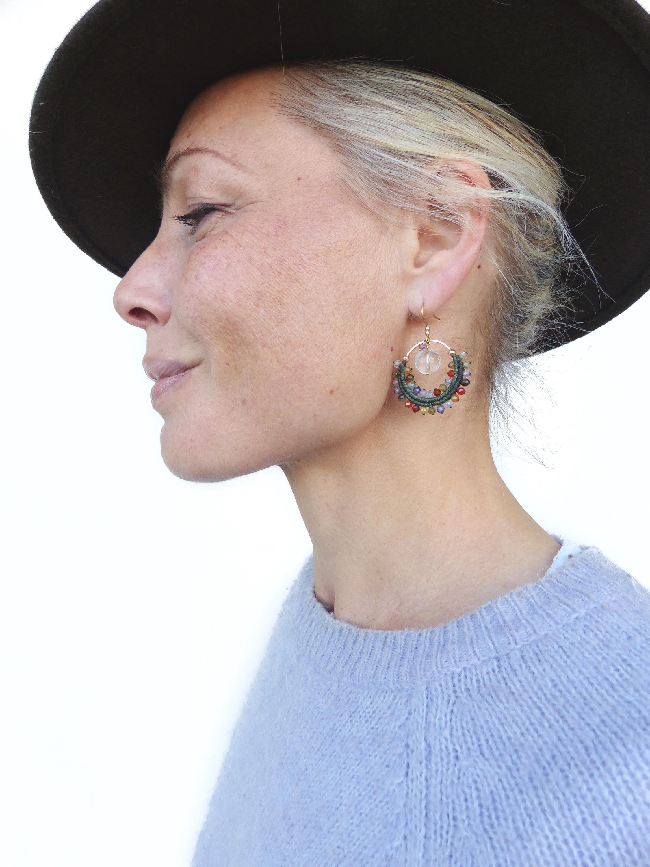 Peut inclure: Boucles d'oreilles créoles dorées avec un cristal transparent et des perles colorées. Les boucles d'oreilles sont attachées à un crochet doré. La femme porte un pull bleu clair et un chapeau marron foncé.