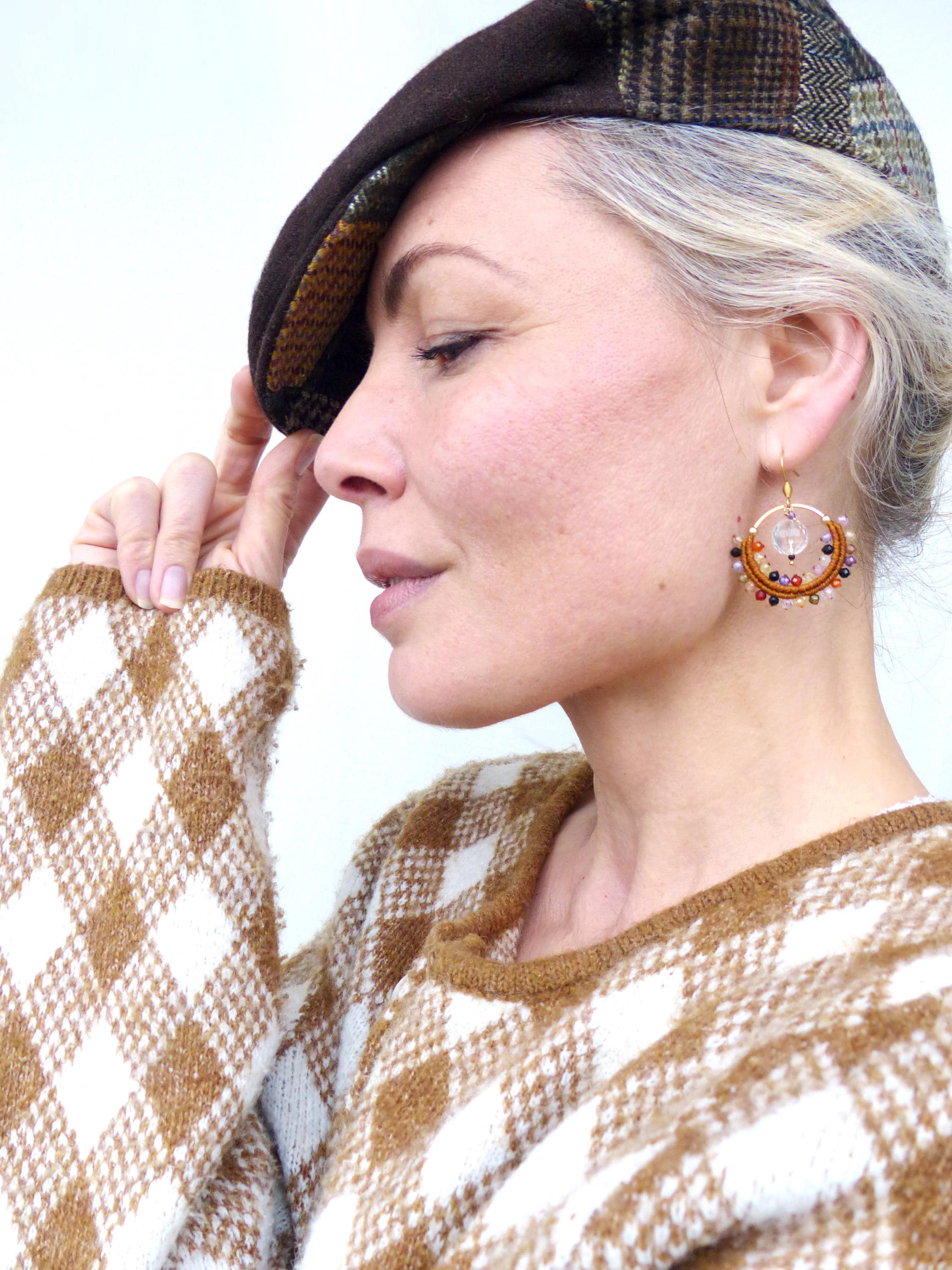 Peut inclure: Une femme porte un béret en tweed marron et un pull à losanges marron et blanc. Elle porte également des boucles d'oreilles dorées avec des perles colorées et un cristal transparent. Les boucles d'oreilles sont le point central de l'image.