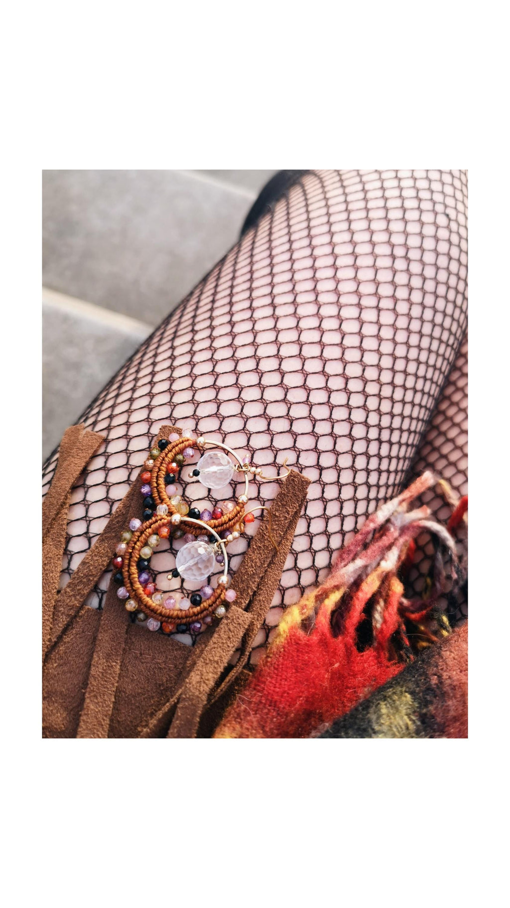 Peut inclure: Boucles d'oreilles créoles faites à la main de style bohème. Les boucles d'oreilles présentent des perles marron, orange, violettes et transparentes, avec un cristal transparent au centre. Elles sont présentées sur un fond de bas résille et de daim marron.