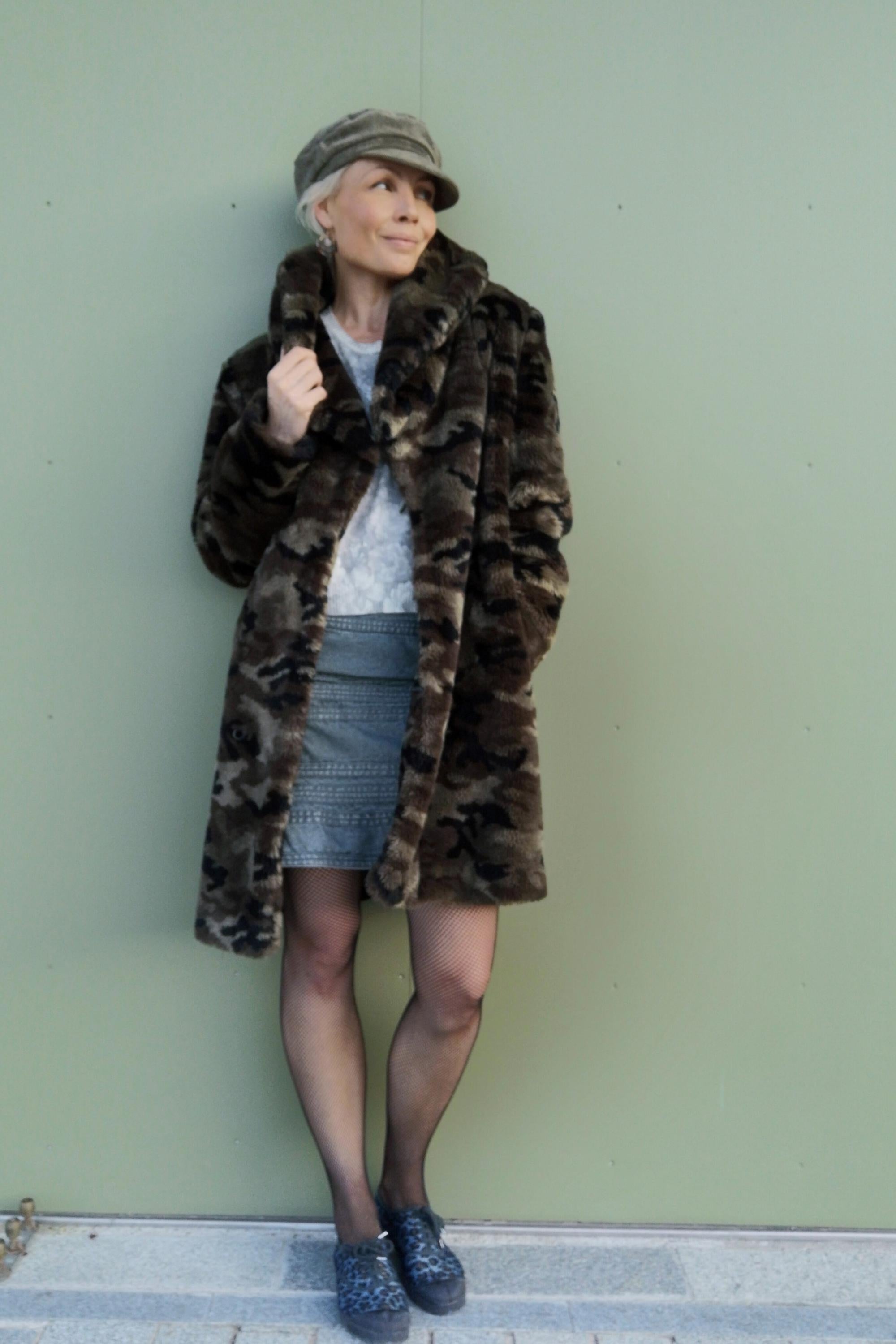 Peut inclure: Une personne porte un manteau en fausse fourrure à motif camouflage, une jupe en jean et une casquette grise. Le manteau est long et a un large col. Des bas résille et des chaussures à motifs complètent la tenue. Le fond est vert clair uni.