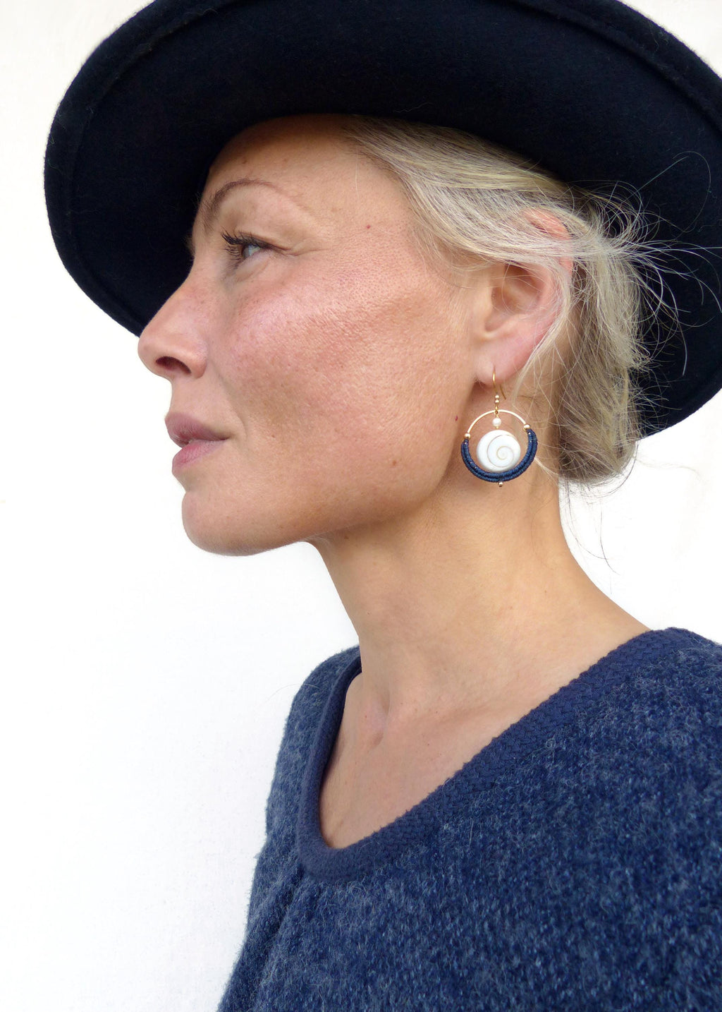 Peut inclure: Une femme porte des boucles d'oreilles dorées avec un motif en spirale blanc et des accents bleu marine. Les boucles d'oreilles sont assorties à un pull bleu et un chapeau noir. La femme a les cheveux blonds et la peau claire.