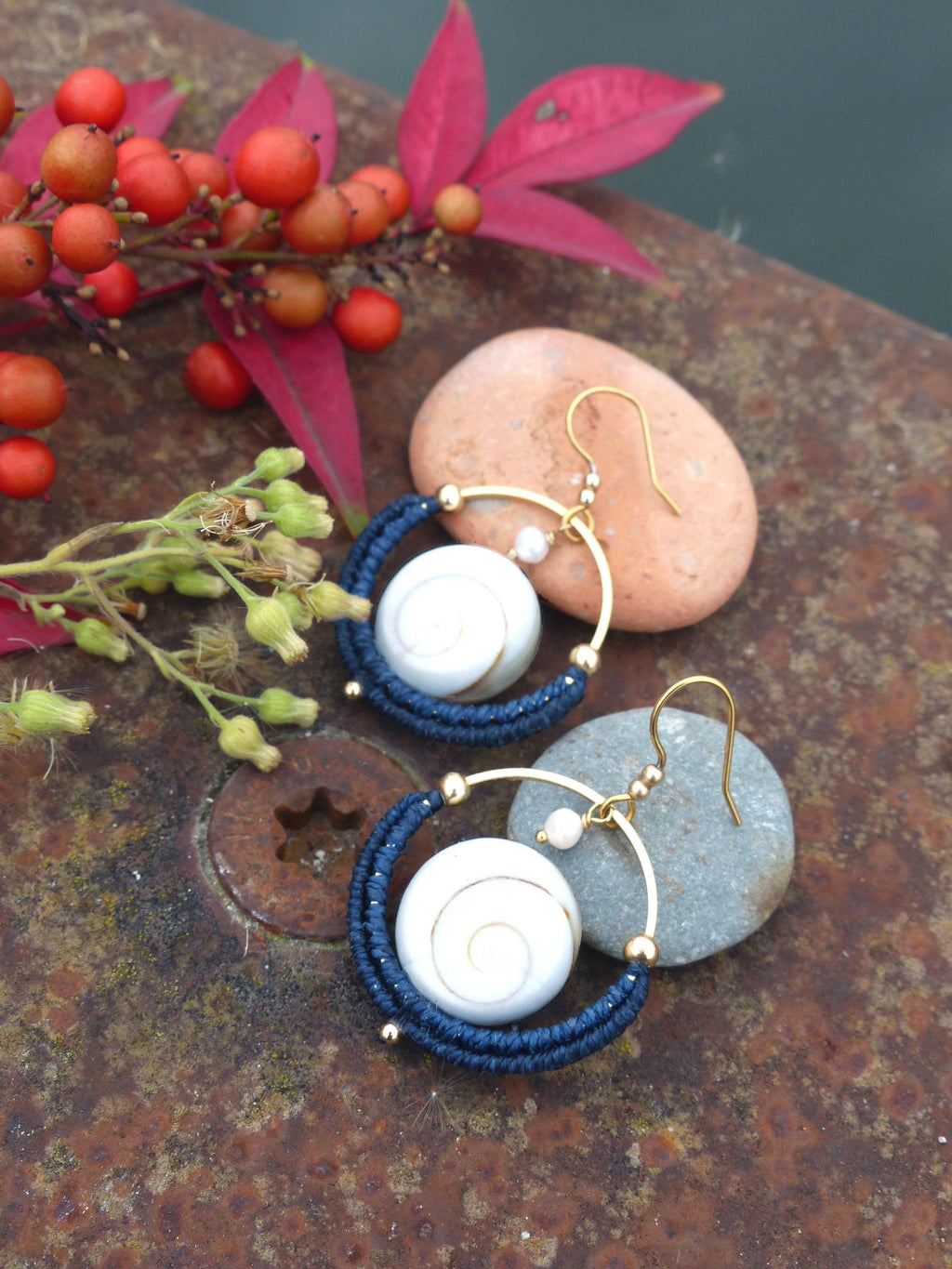 Peut inclure: Boucles d'oreilles artisanales avec un motif en spirale blanc dans un cercle en macramé bleu marine. Les boucles d'oreilles ont des crochets et des accents dorés, avec une petite perle. Elles sont présentées sur une surface rouillée texturée.