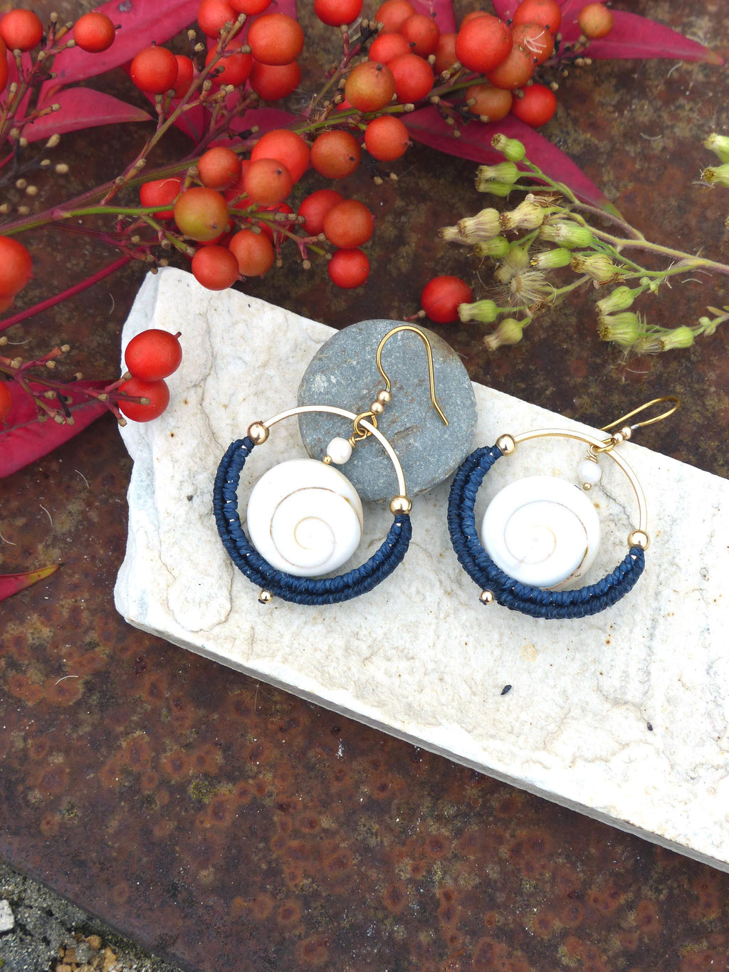 Peut inclure: Boucles d'oreilles faites à la main avec des cerceaux dorés, des spirales blanches et des détails en fil bleu marine. Les boucles d'oreilles sont présentées sur une surface en pierre blanche, avec des baies rouges et du feuillage en arrière-plan.